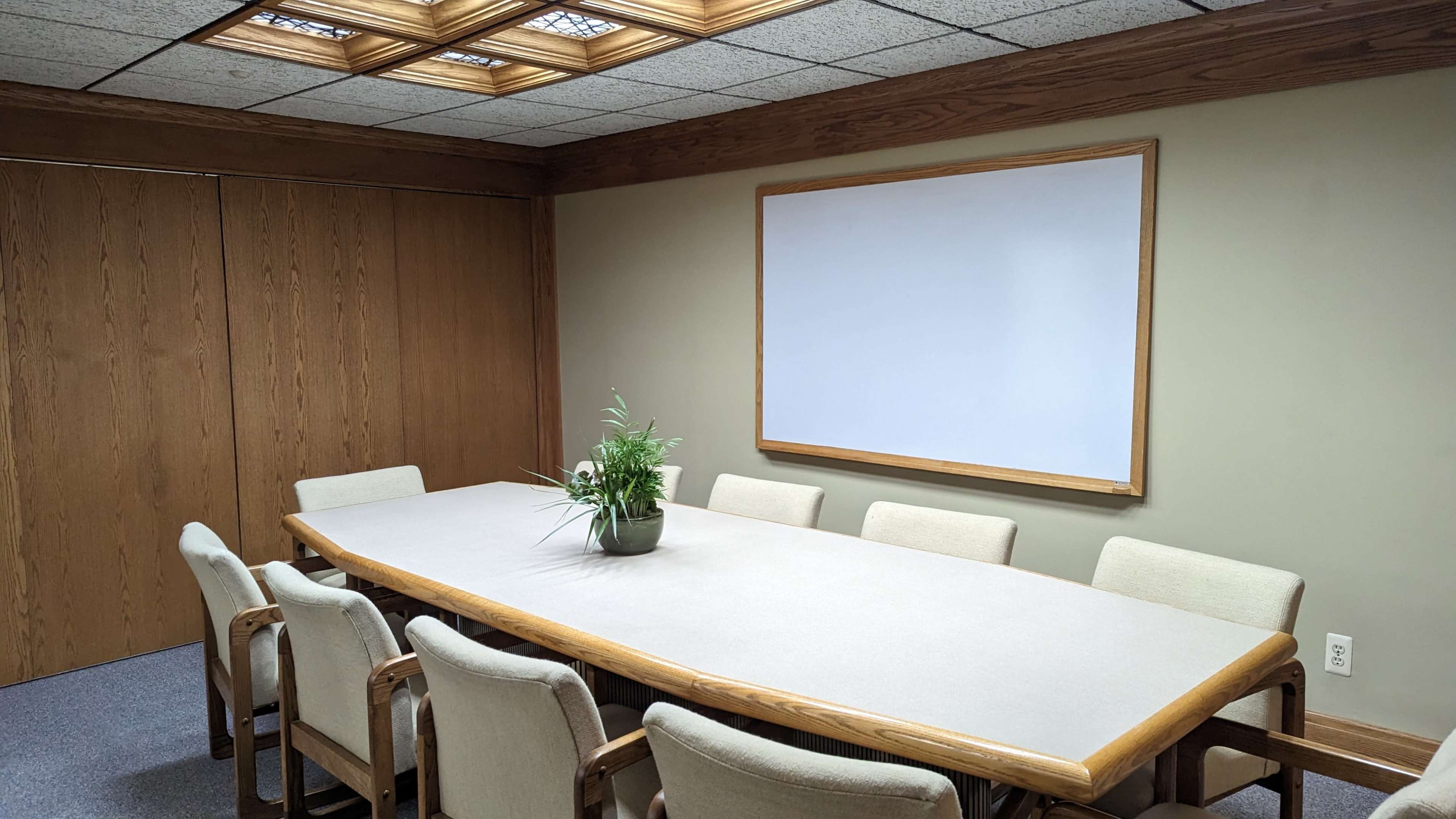 The image shows a conference room with a long table surrounded by chairs, a potted plant in the center, and a whiteboard on the wall.
