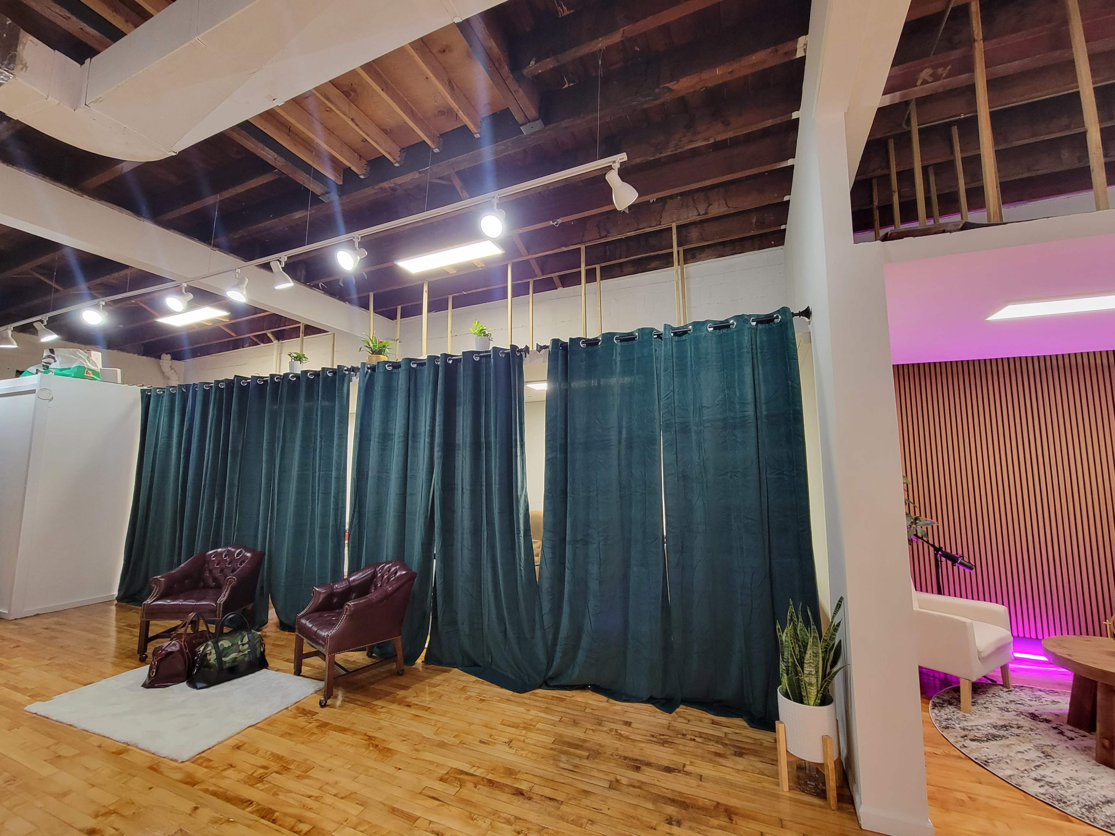 The image shows a room with wooden floors featuring two burgundy chairs, green curtains, and plants, alongside a section with pink lighting and a minimalist setup.