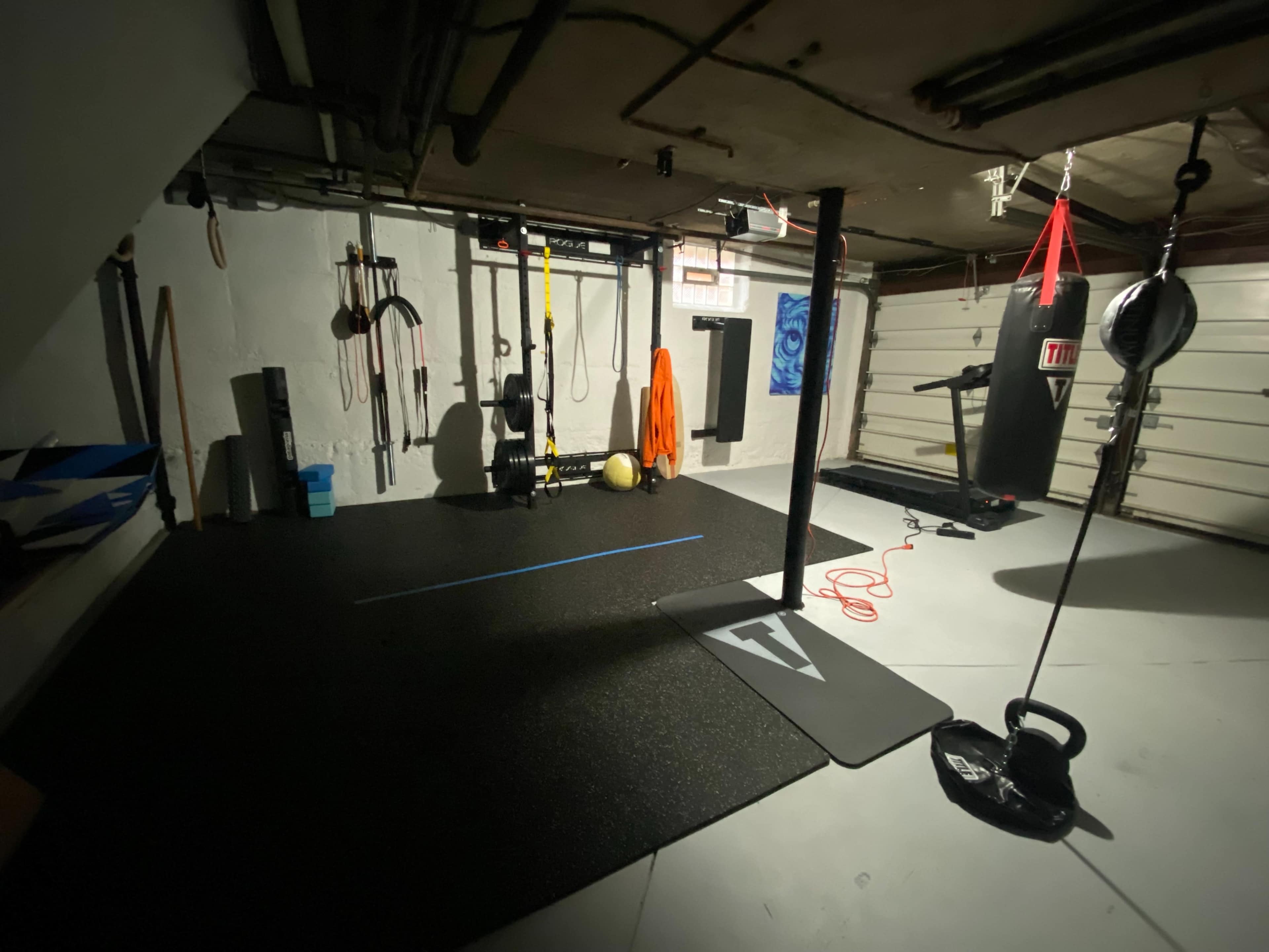 A dimly lit home gym featuring a punching bag, kettlebell, exercise mats, and various workout equipment hanging on the walls.
