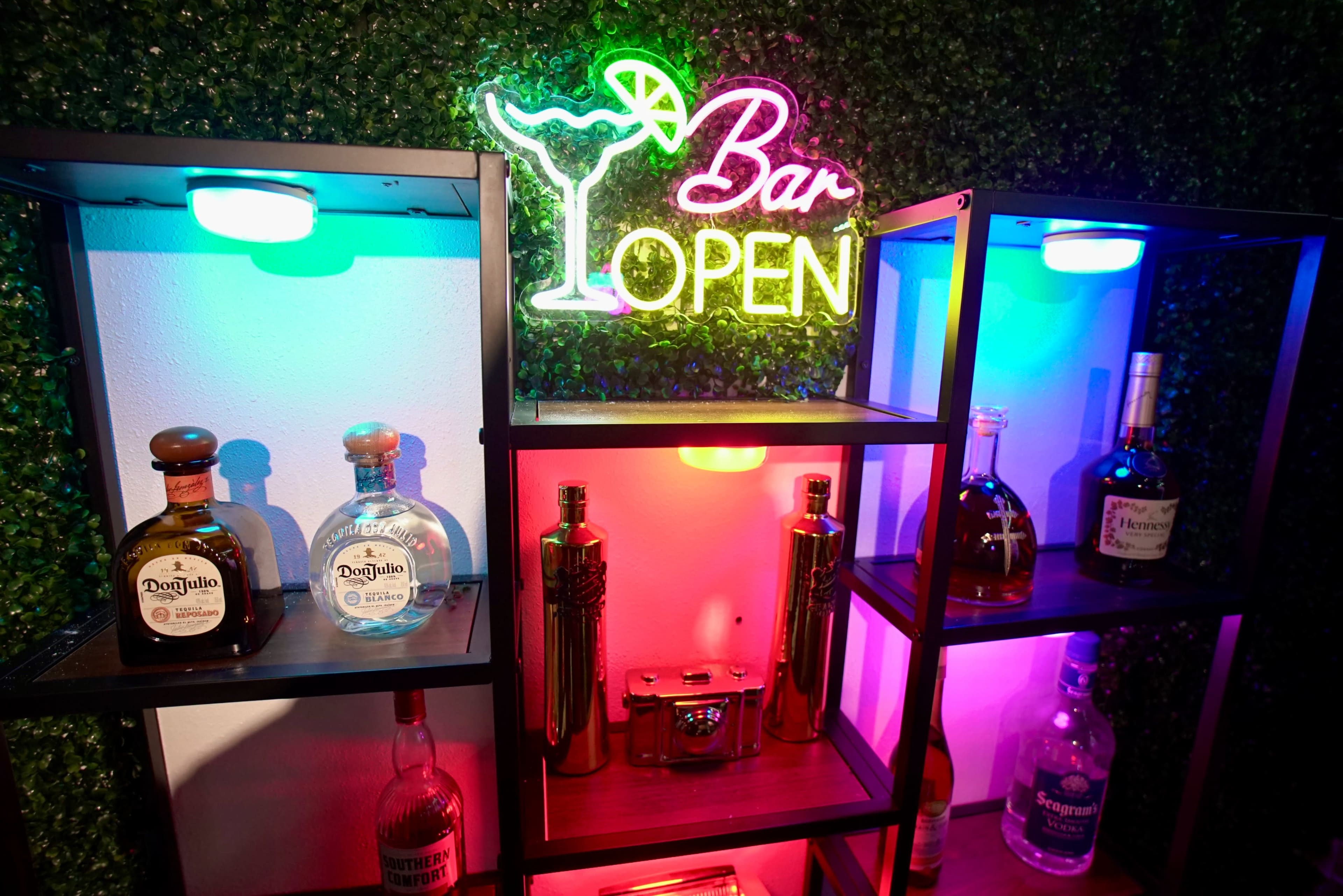 The image shows a backlit shelf displaying various bottles of alcohol alongside a neon sign that reads "Bar OPEN."