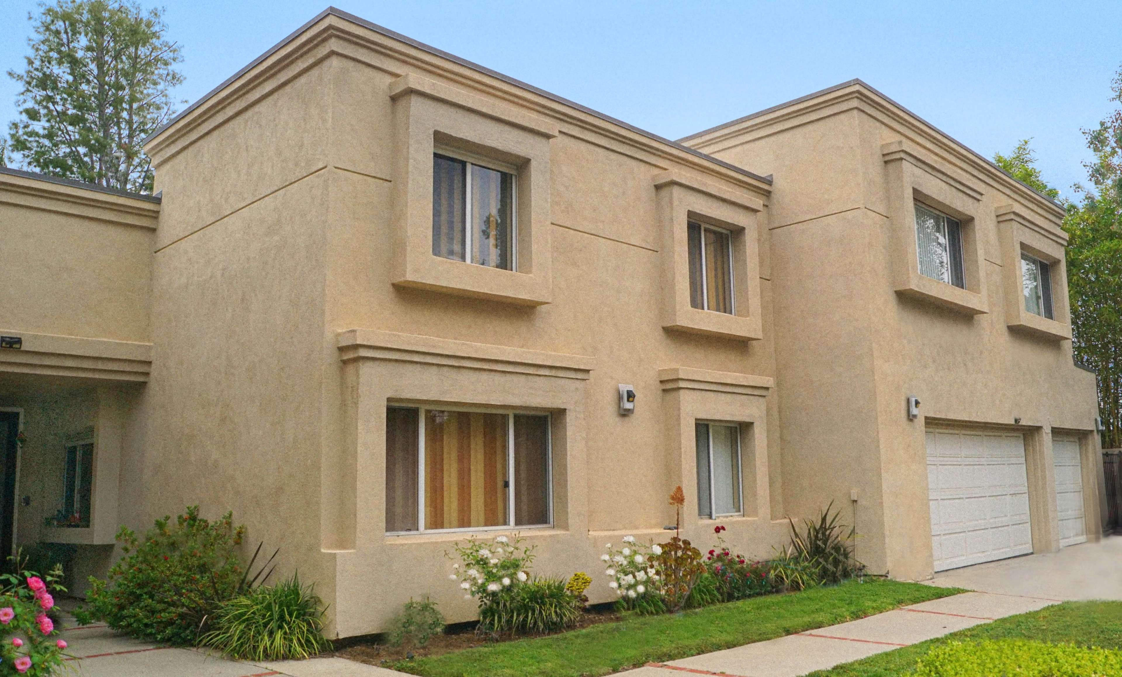 The image shows a two-story residential building with beige stucco exterior, featuring multiple windows and a driveway leading to a two-car garage.
