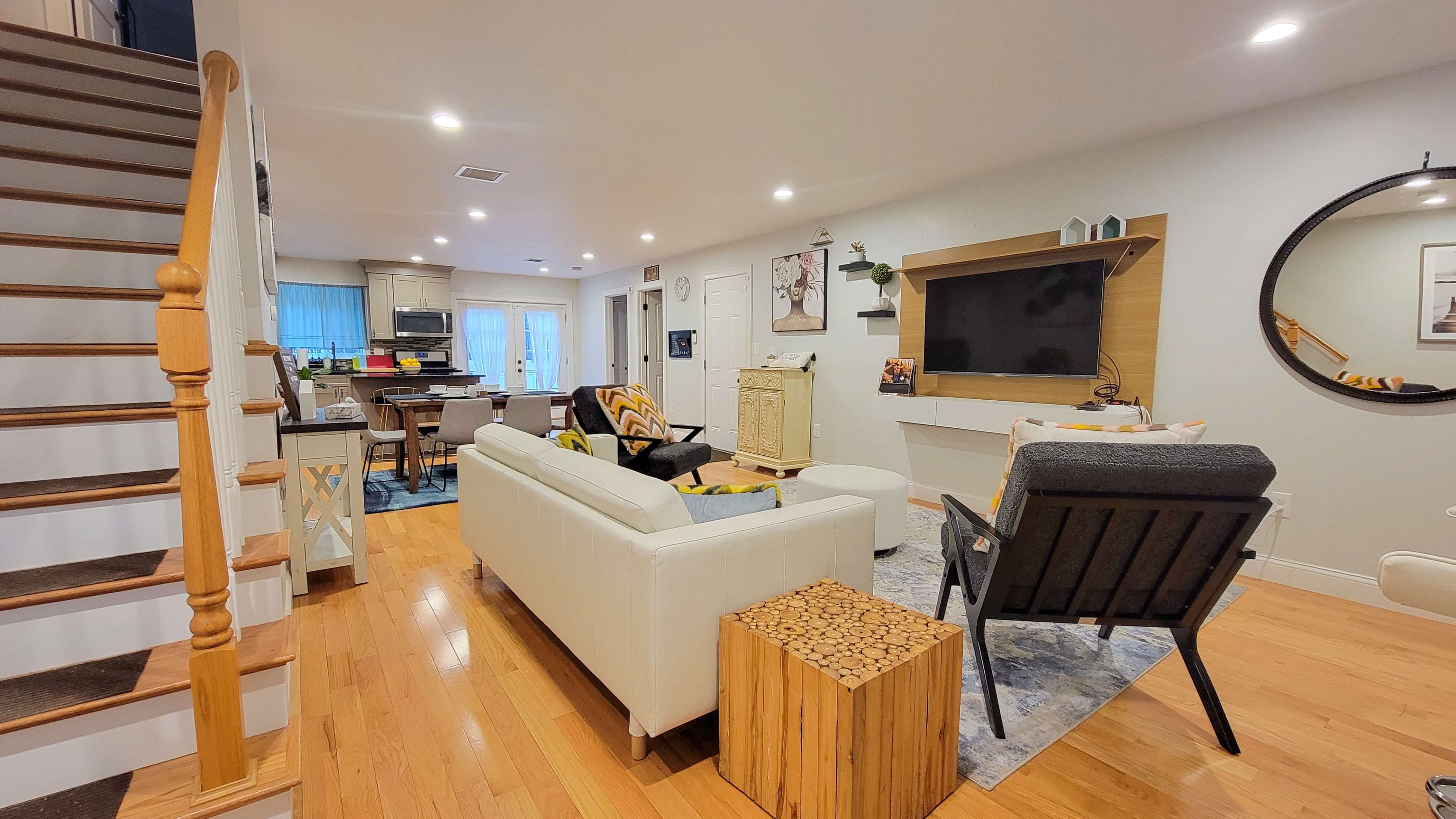 A modern living room with a staircase on the left, a sofa and armchair facing a wall-mounted television, and a dining area visible in the background.