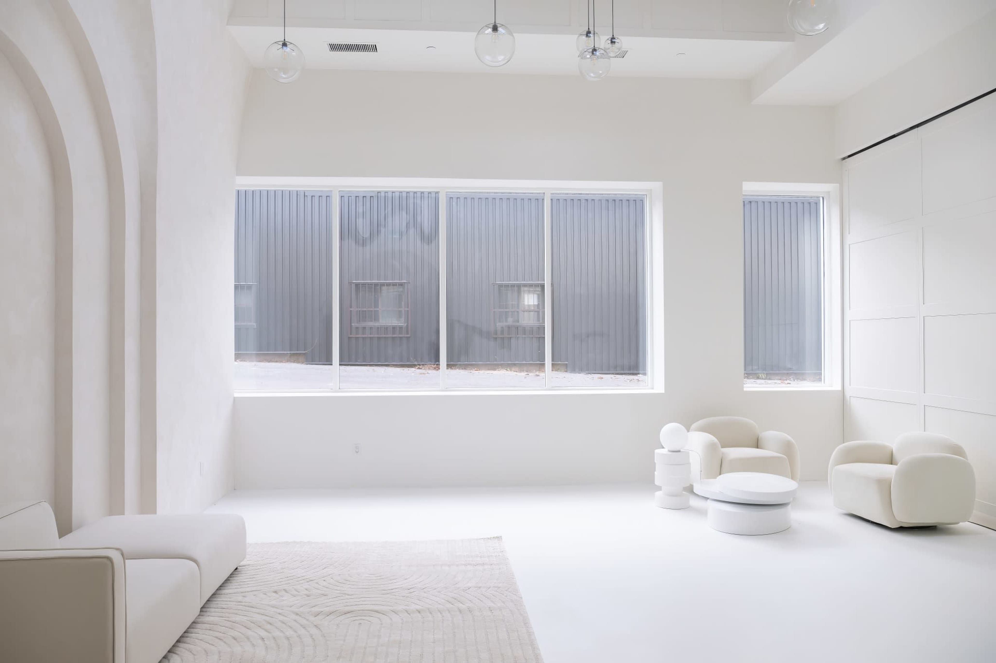 A minimalist, white interior space featuring a sofa, round coffee table, and modern chairs, with large windows revealing a gray exterior wall.