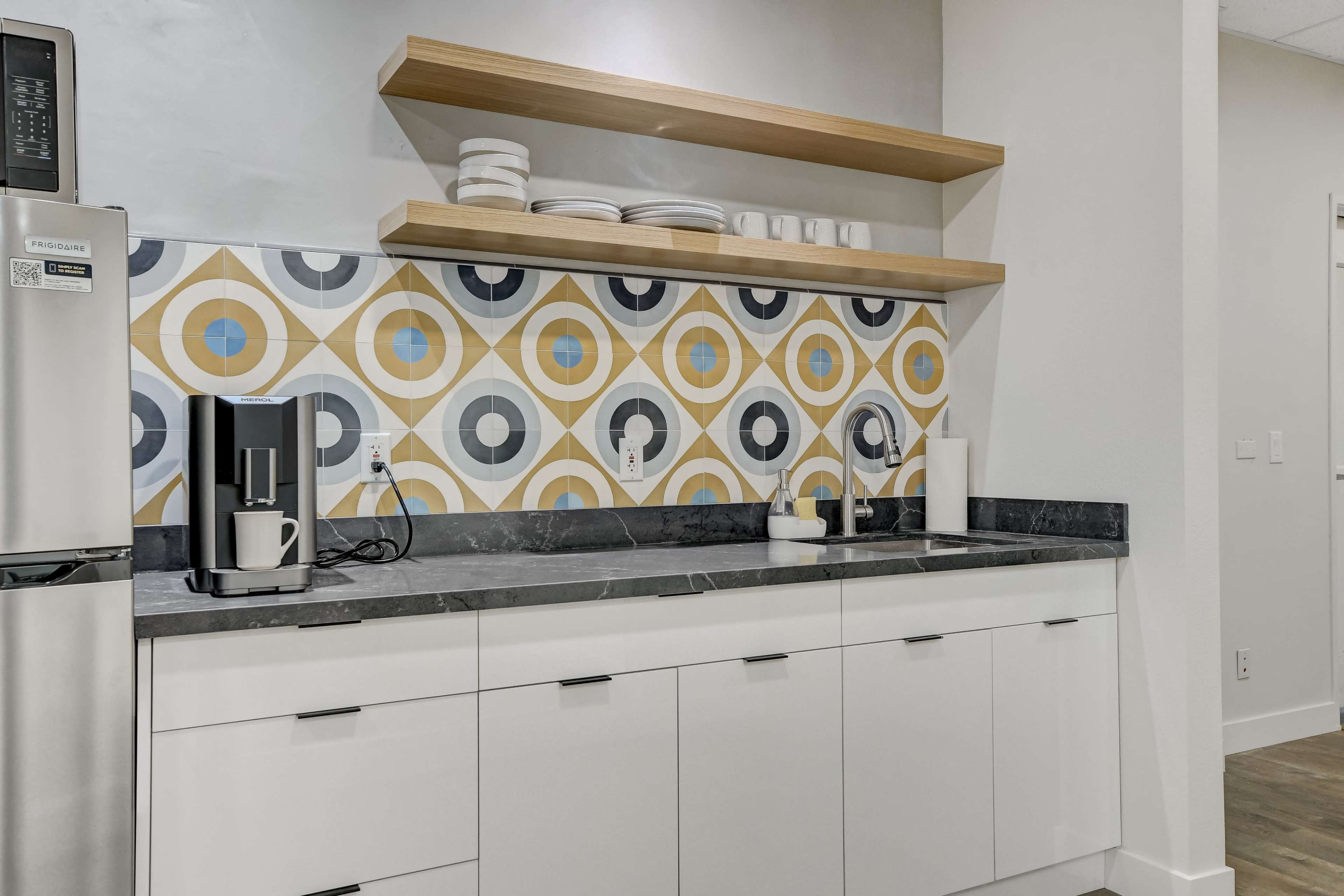 A modern kitchen features a gray countertop with a coffee maker, white dishes on floating shelves, and a colorful geometric tile backsplash.