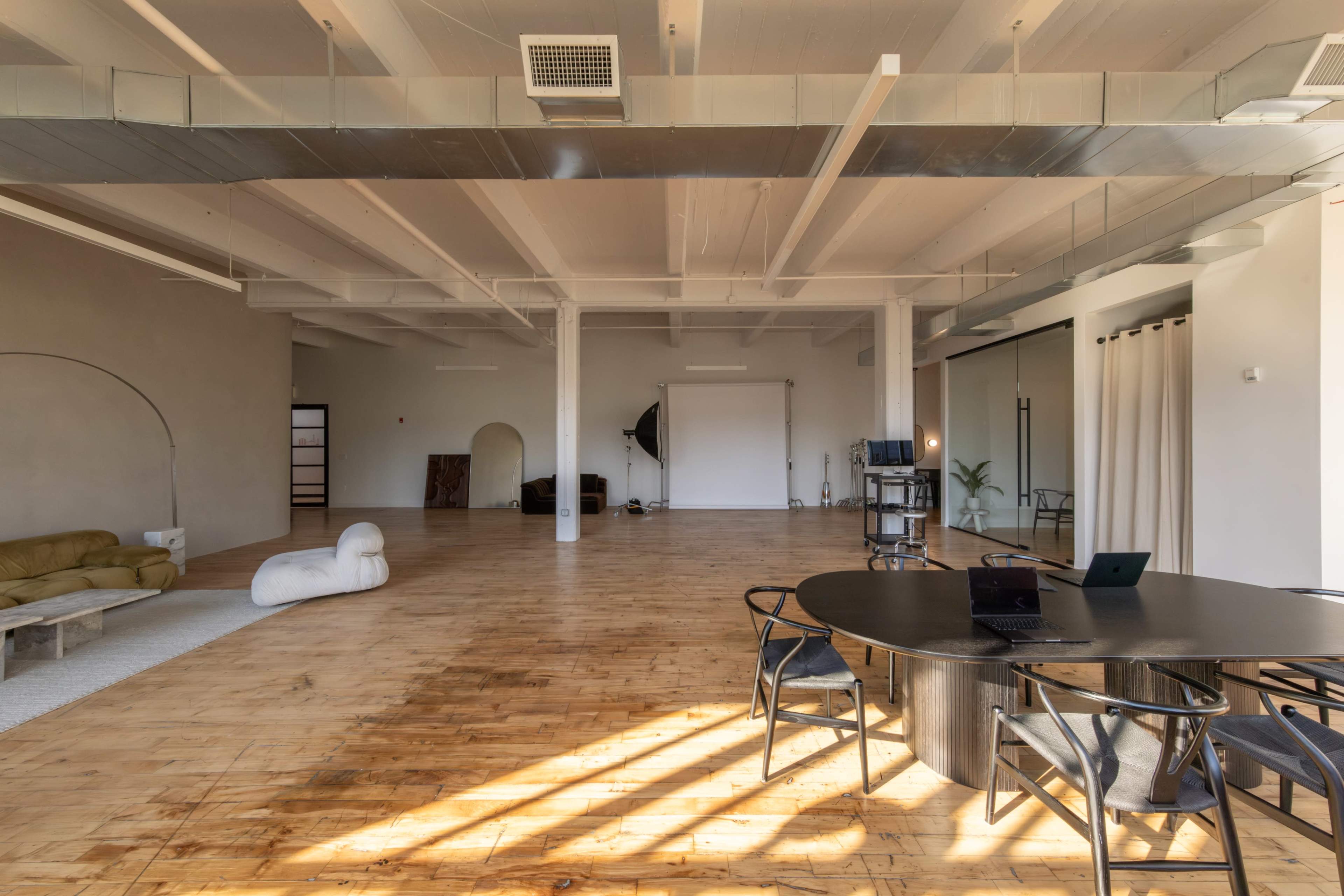 The image shows a spacious, minimalistic interior with a wooden floor, large windows, and various furniture pieces, including a round table and a sectional sofa.