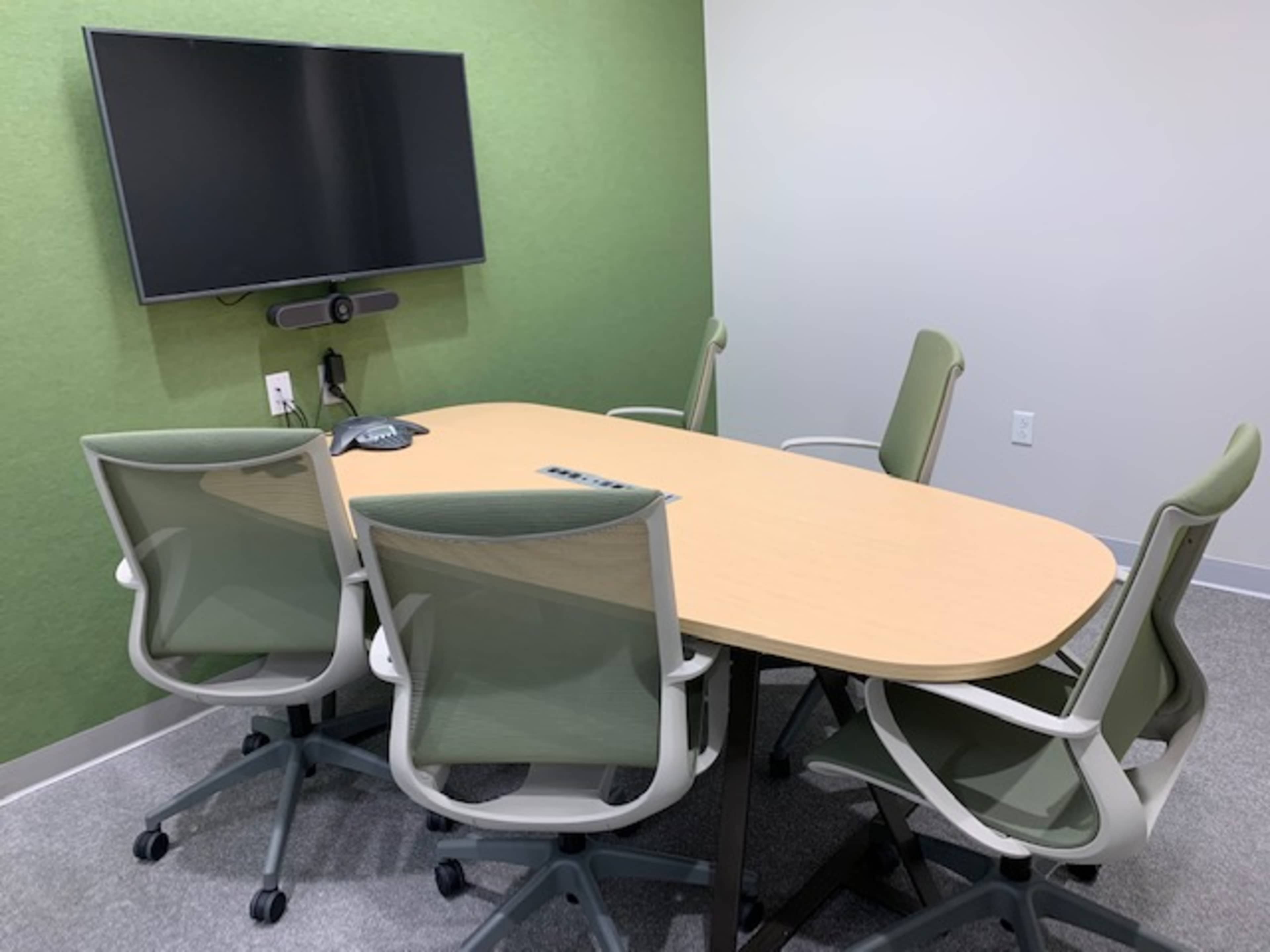 A small meeting room with a wooden oval table, six green office chairs, and a wall-mounted television.