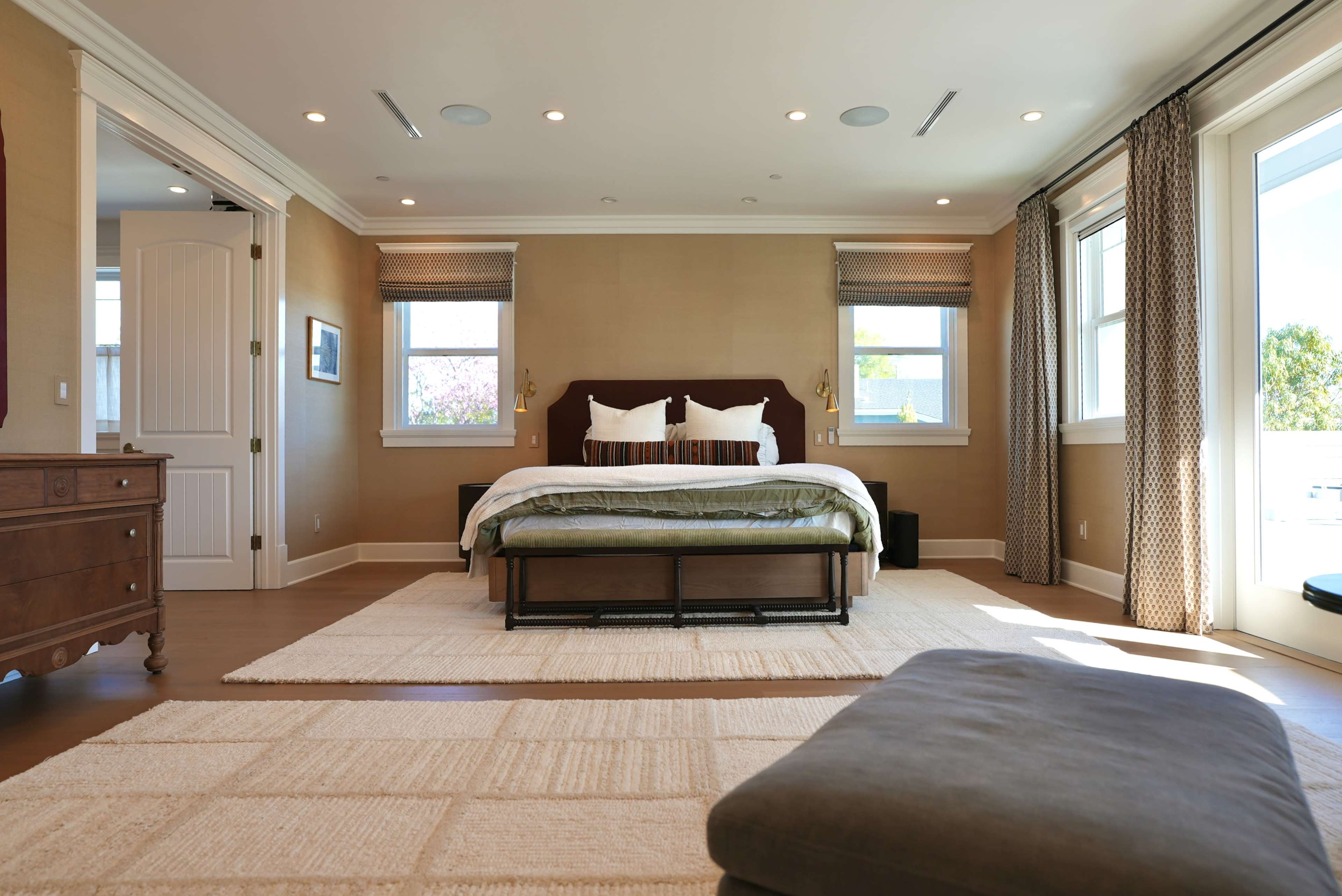 The image shows a well-lit bedroom featuring a large bed, two windows with shades, a wooden dresser, and a beige area rug on the hardwood floor.