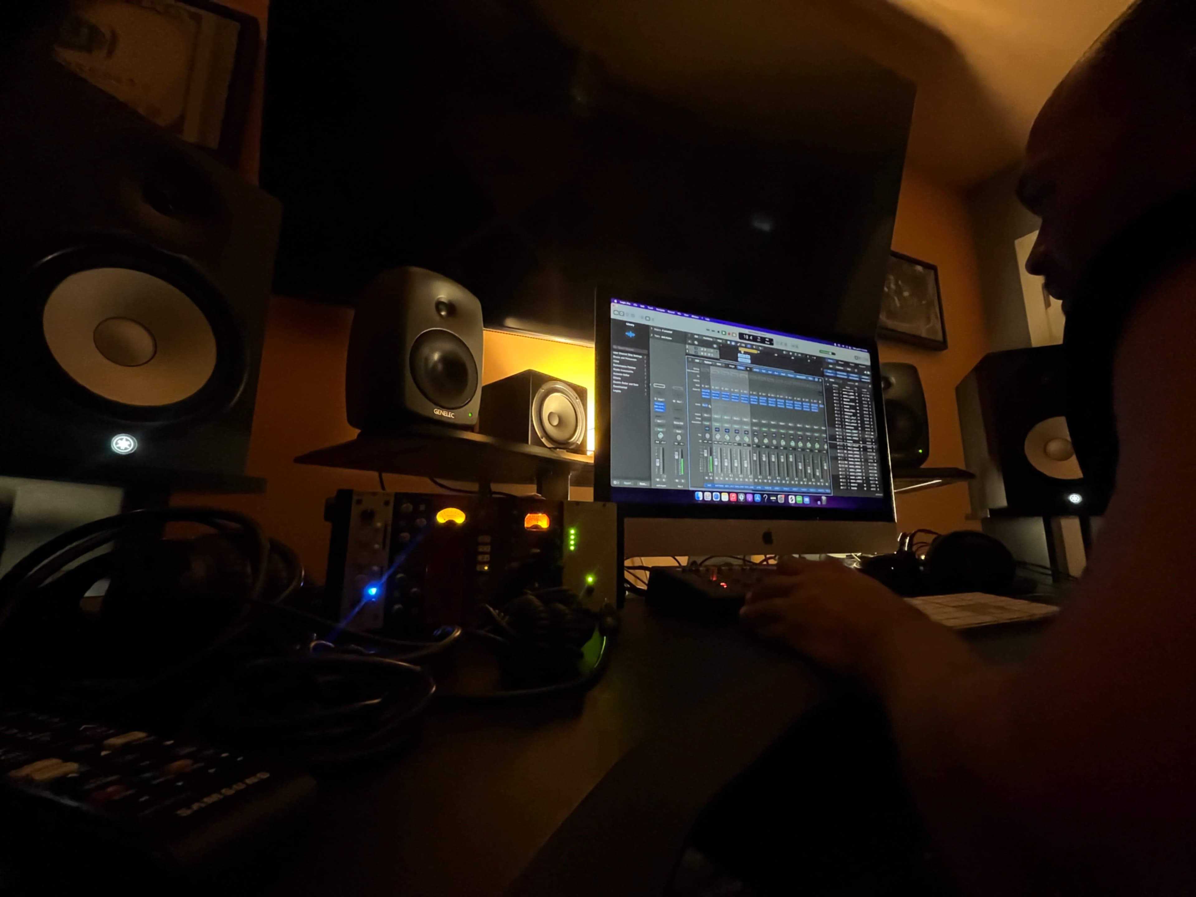 A person is working at a music production setup with multiple speakers, a computer displaying audio software, and various cables and equipment on the desk.
