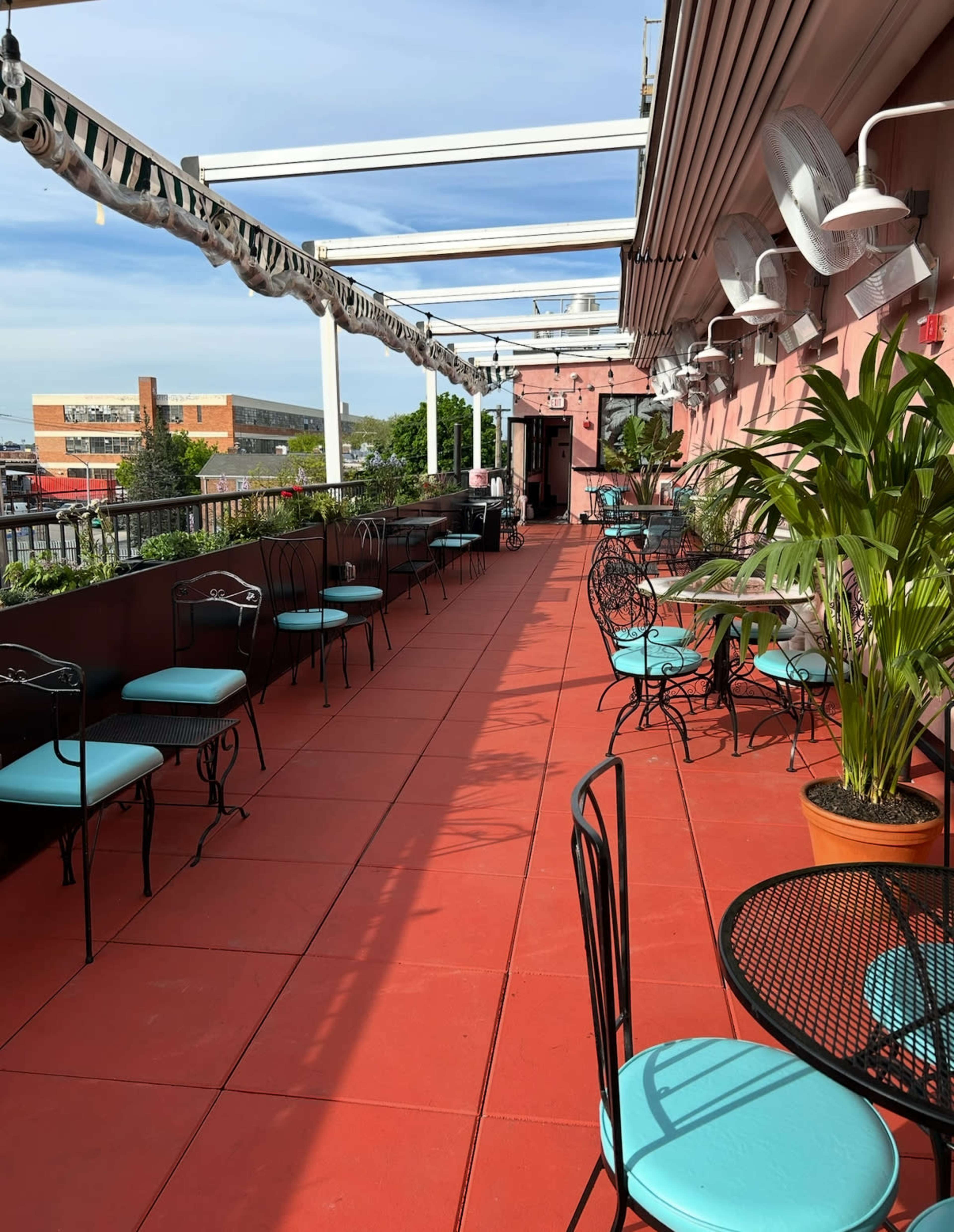 A rooftop space featuring empty tables and chairs with turquoise seats, surrounded by potted plants and shaded by a canopy.