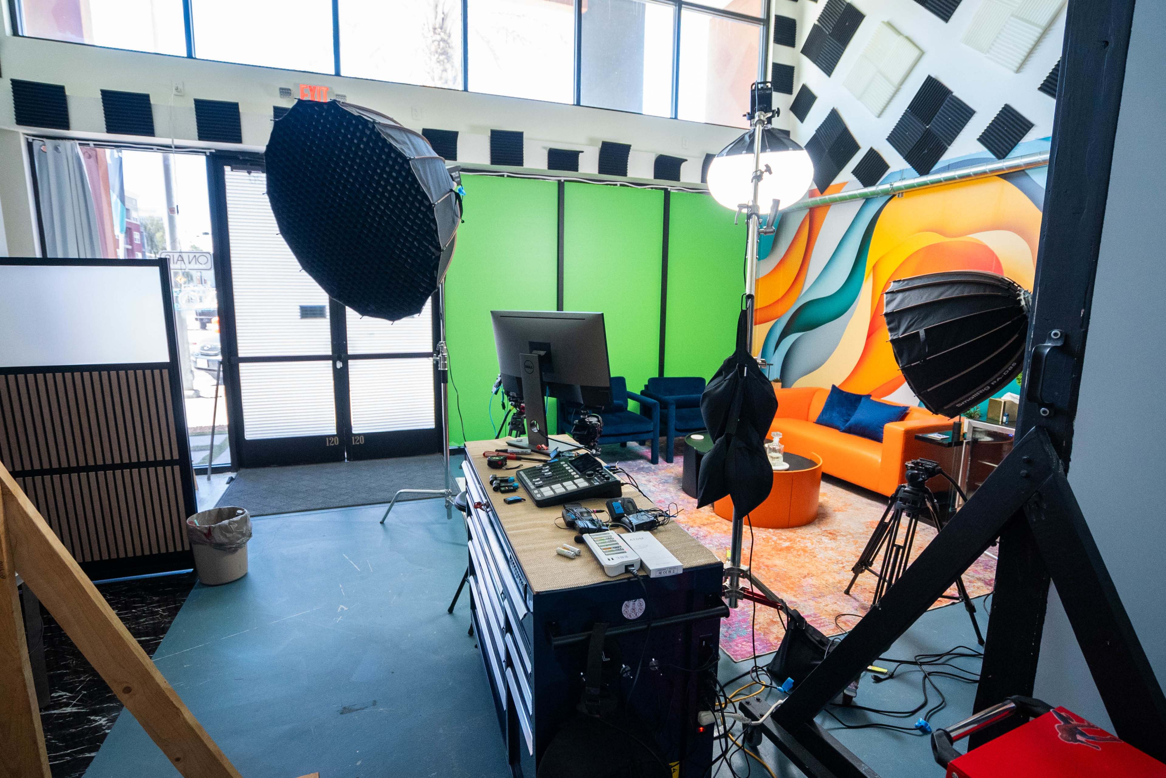 The image shows a brightly lit studio space equipped with various lighting setups, a green backdrop, a computer workstation, and colorful furnishings.