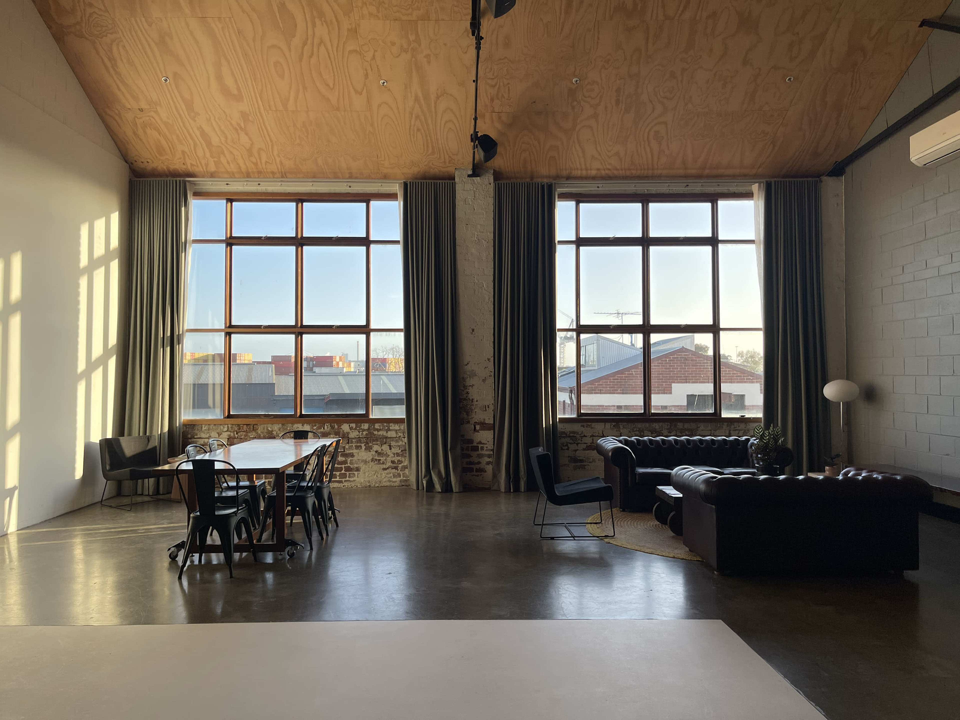 The image shows a spacious industrial-style living area with large windows, a wooden ceiling, a dining table, and a leather couch.