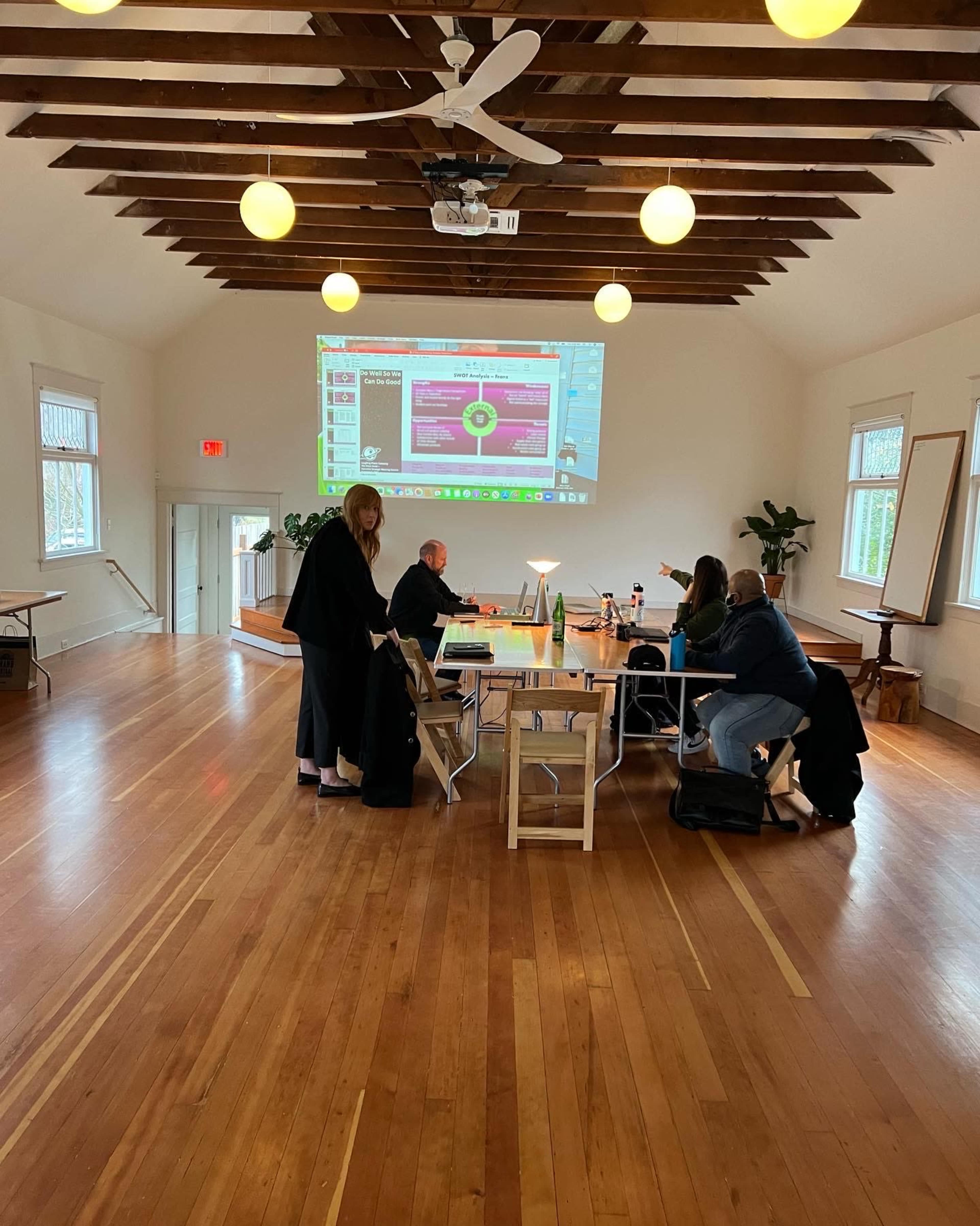 A group of people is gathered around tables in a spacious room with wooden floors and a large screen displaying a presentation.