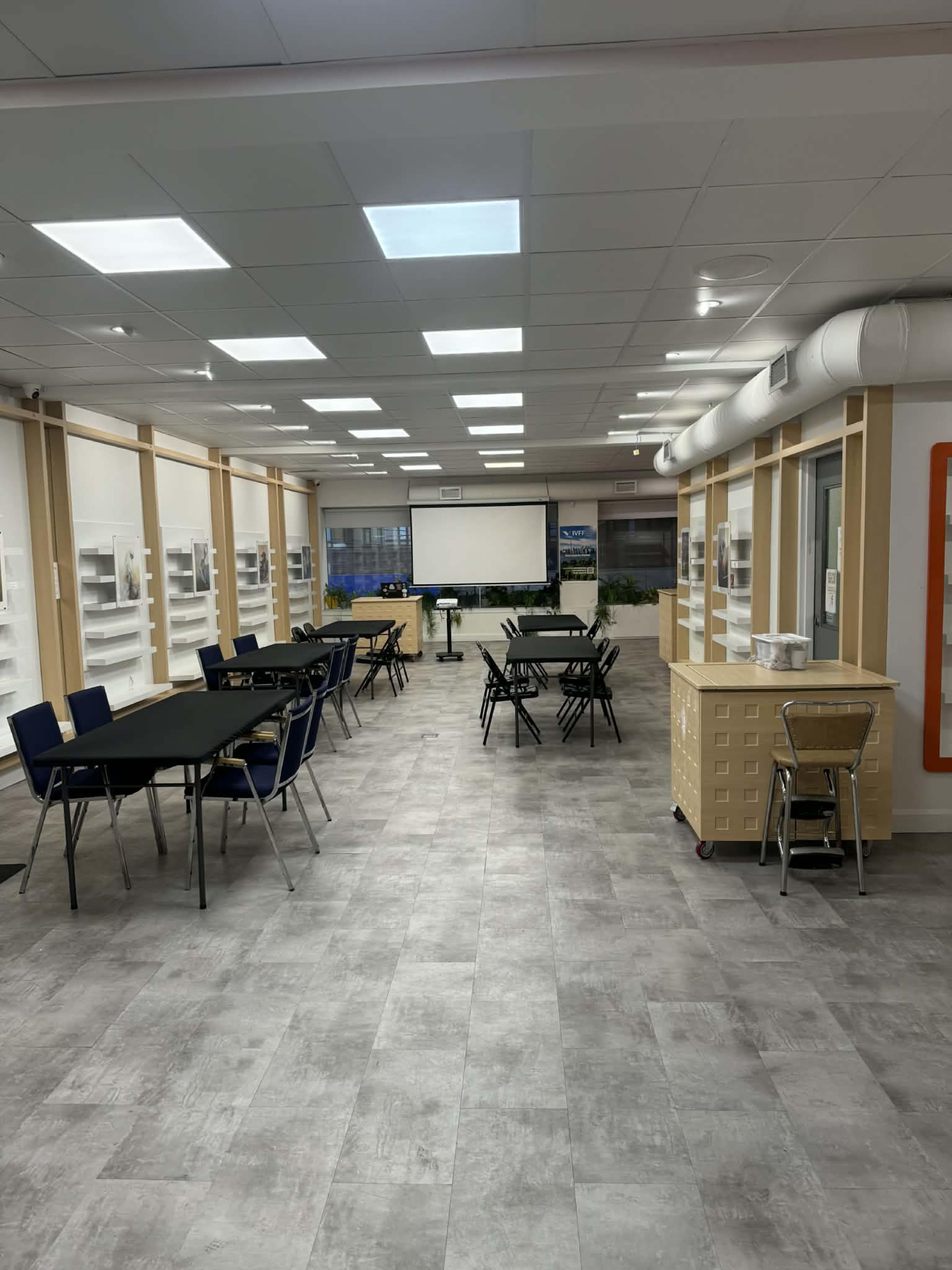 The image shows a spacious, well-lit classroom with tables and chairs arranged for small group work, a presentation screen at the front, and storage units along the walls.