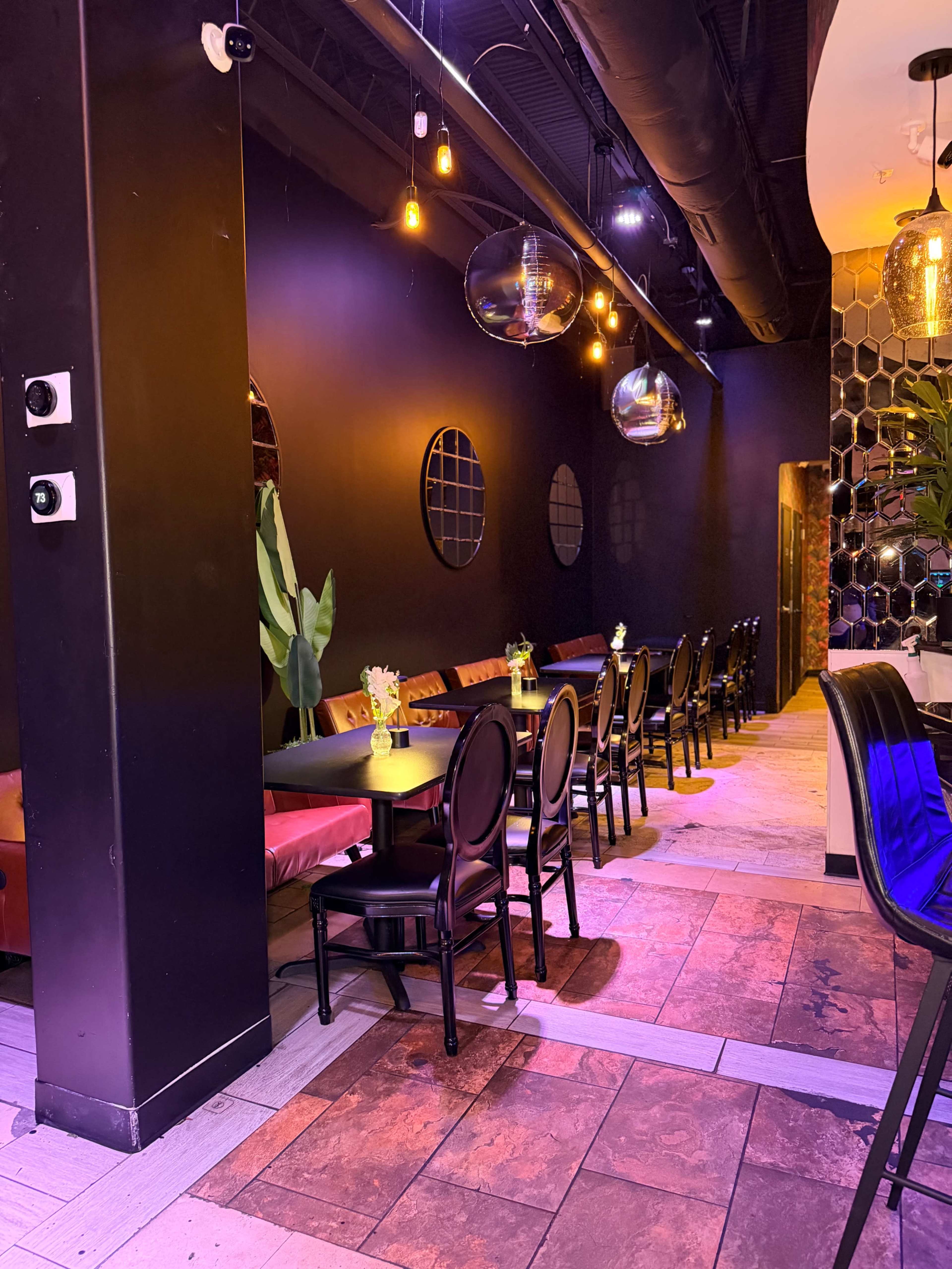The image shows a modern restaurant interior featuring dark walls, pendant lighting, and rows of black dining chairs at tables, along with decorative plants and wall mirrors.