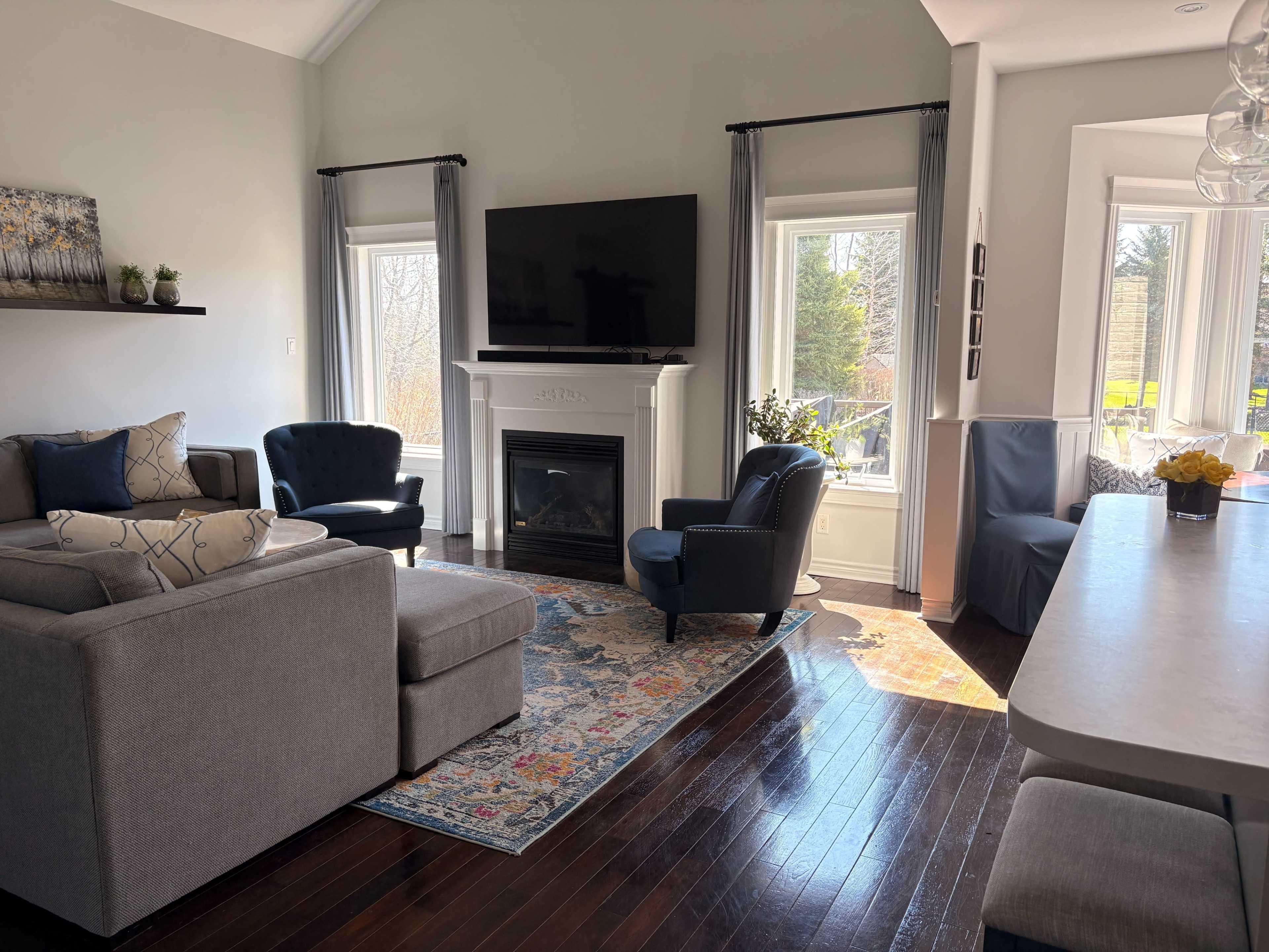 The image shows a modern living room featuring a sectional sofa, two armchairs, a fireplace, and large windows with natural light illuminating the space.