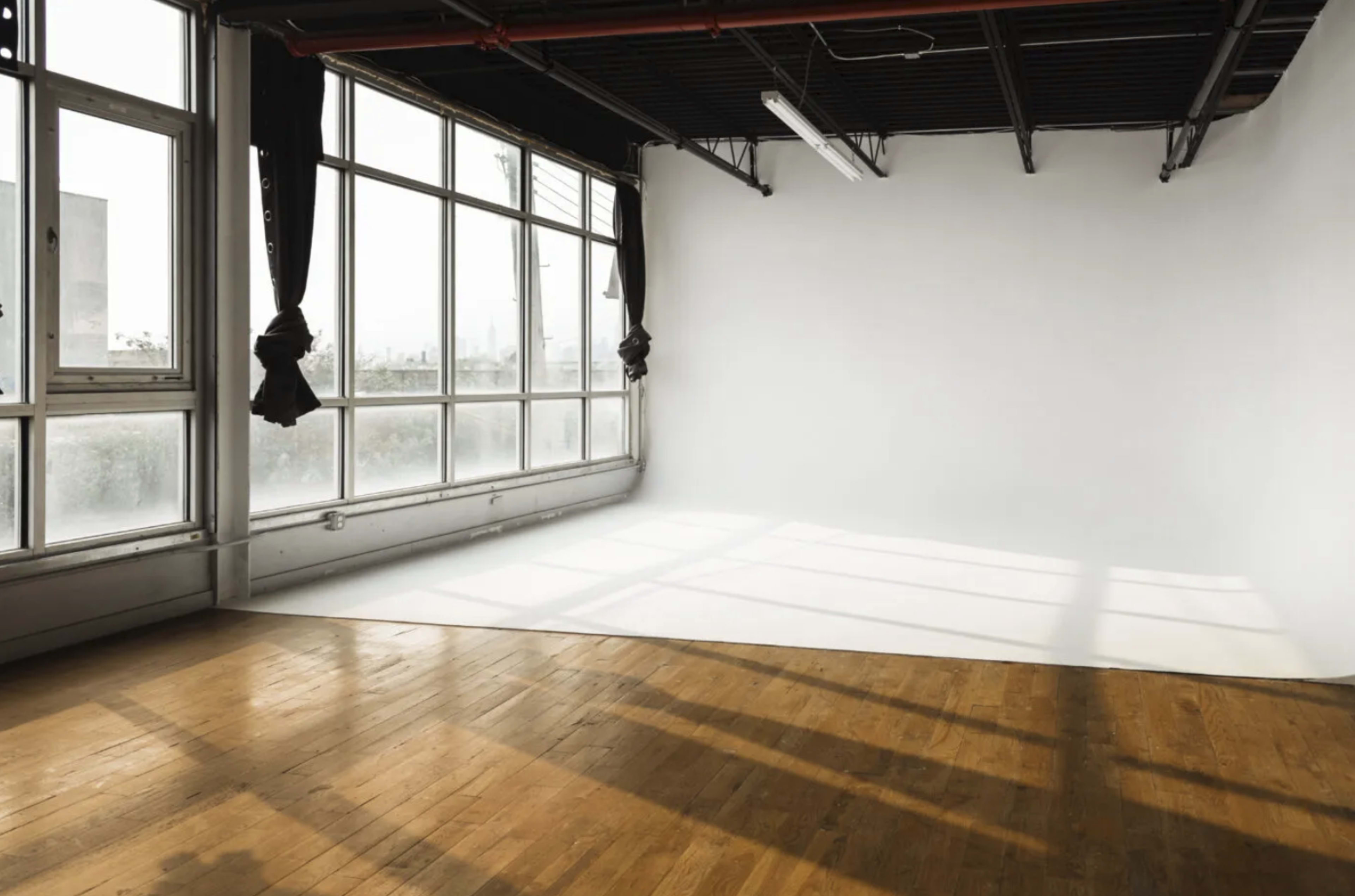 A spacious room with large windows, white walls, and a wooden floor, featuring a seamless white backdrop and natural light casting shadows.