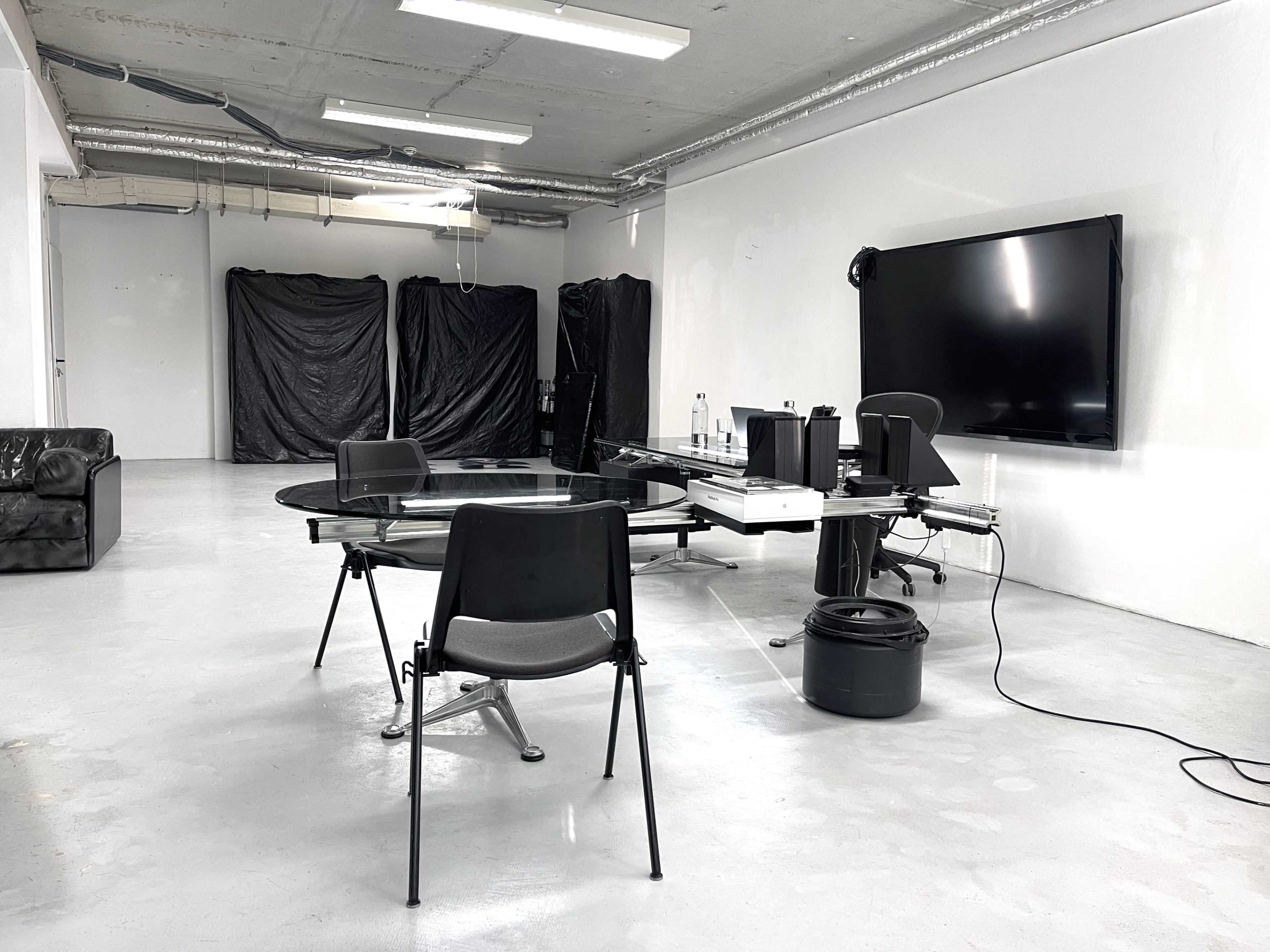 The image shows a minimalist office space with a glass table, black chairs, and a large screen mounted on the wall.