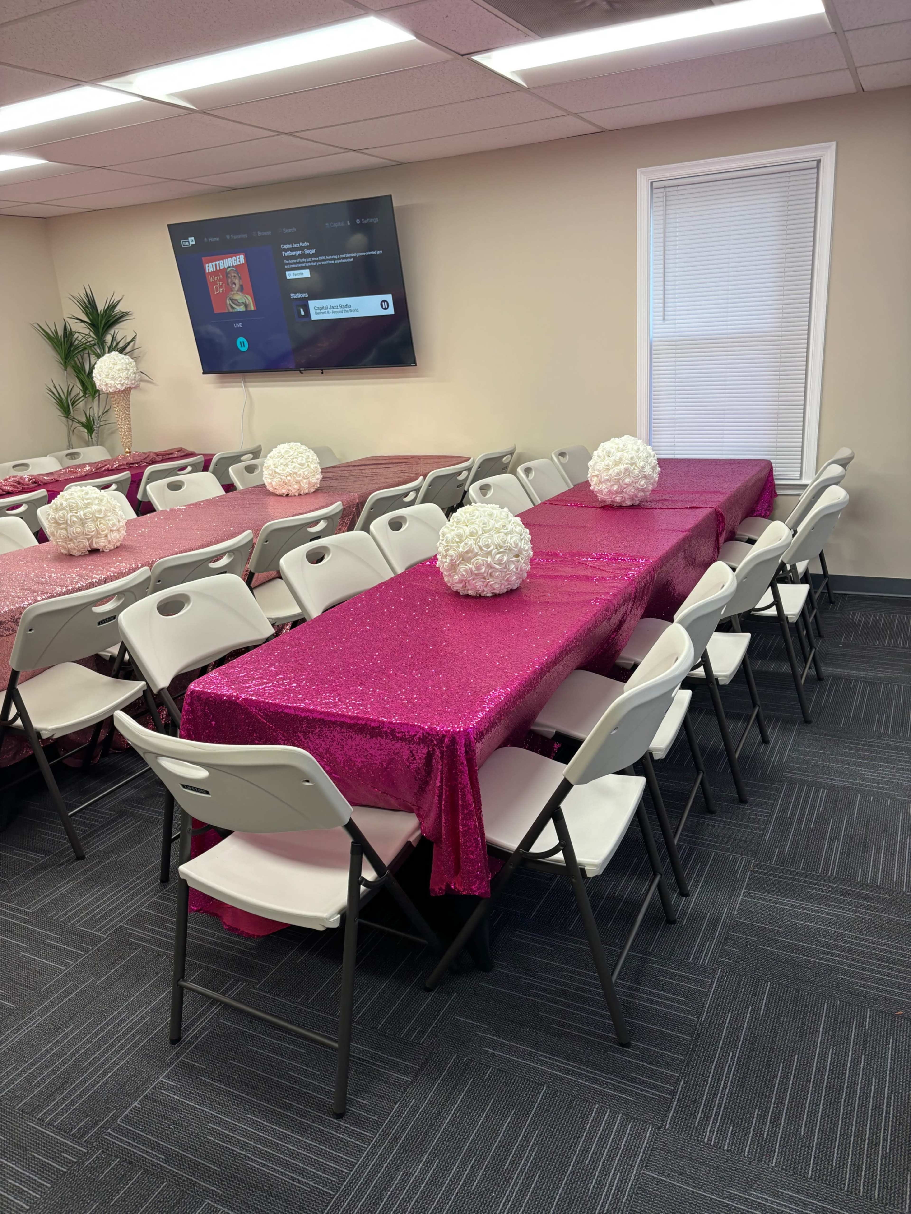 A room with pink sequin tablecloths covering several tables arranged in rows, each adorned with a floral centerpiece, and equipped with white folding chairs.