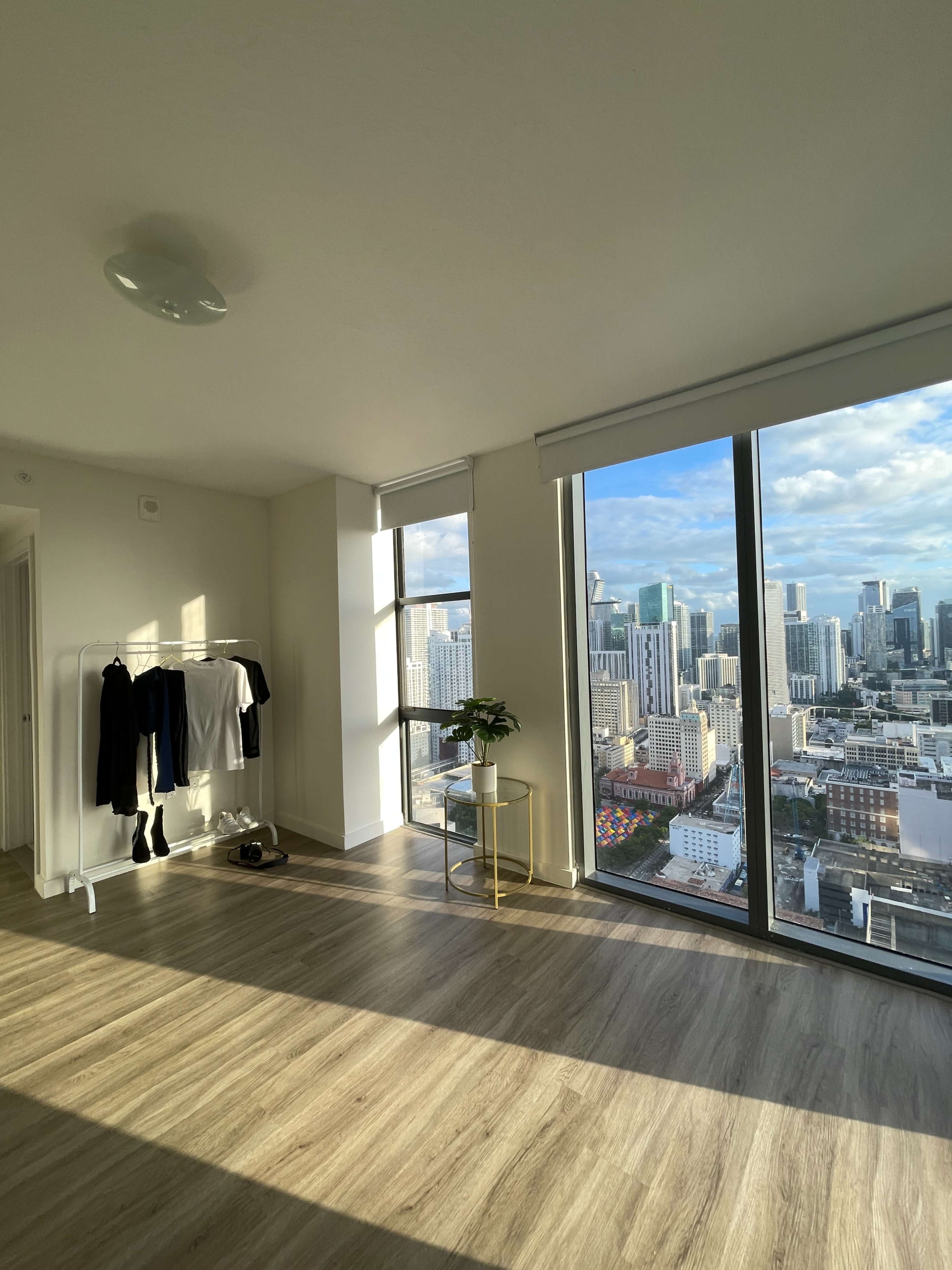 A room with large windows showing a cityscape, featuring a clothing rack, a small plant on a table, and sunlight streaming across the floor.