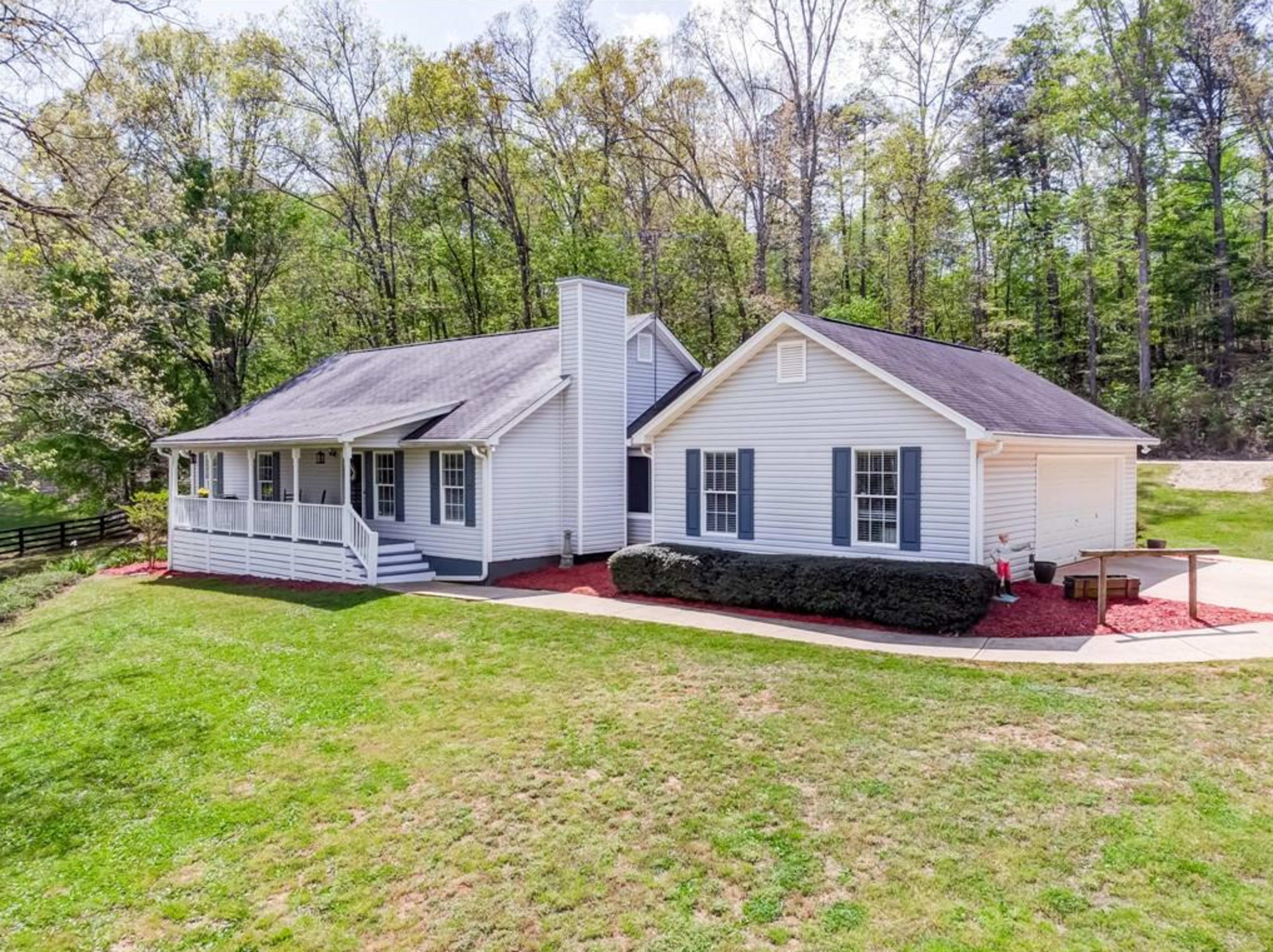 A single-story house with a covered porch and attached garage is situated on a green lawn surrounded by trees.