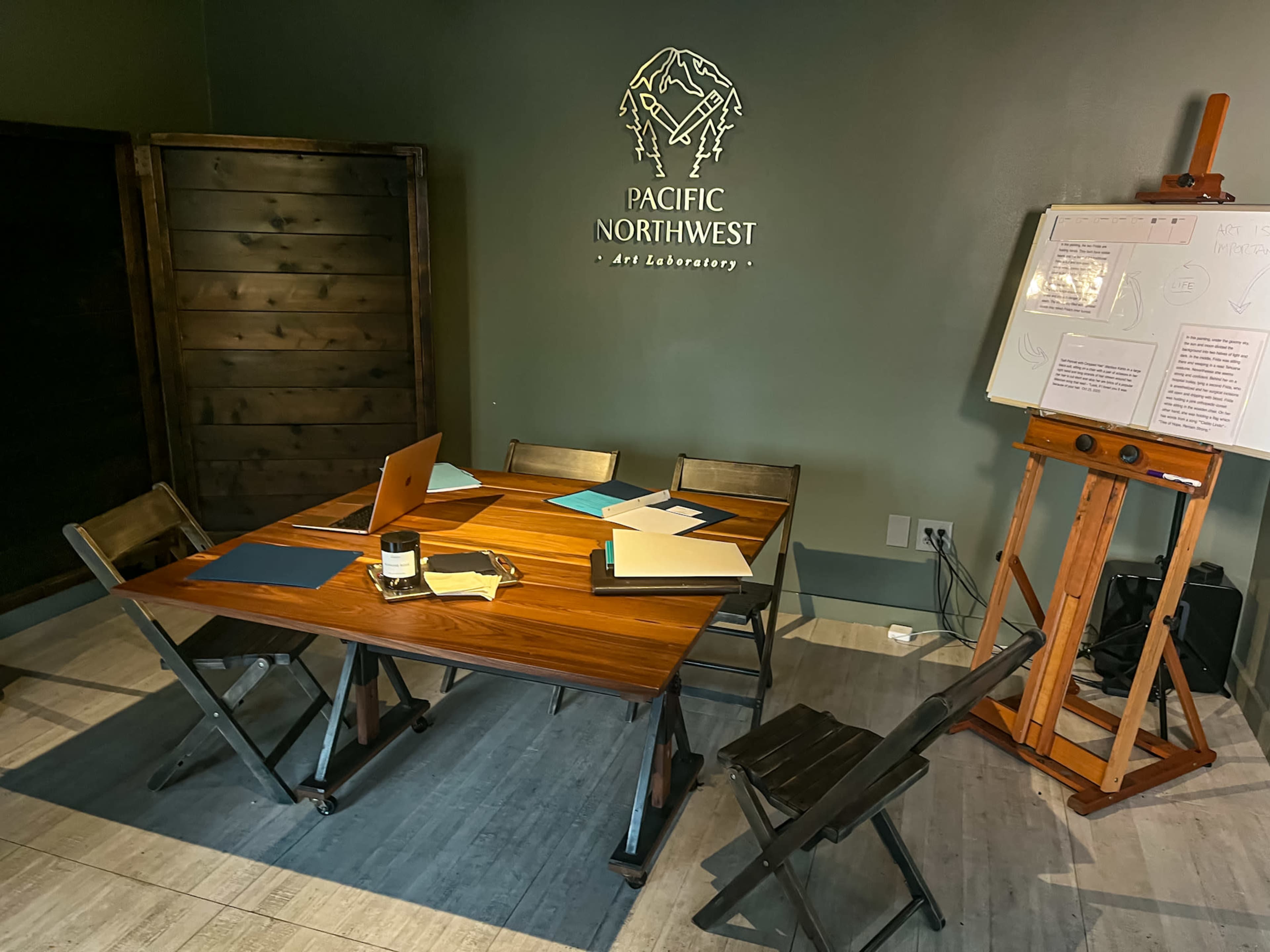 A wooden table with chairs is set up in a room featuring a wall sign that reads "PACIFIC NORTHWEST - Art Laboratory," alongside an easel and various art supplies.