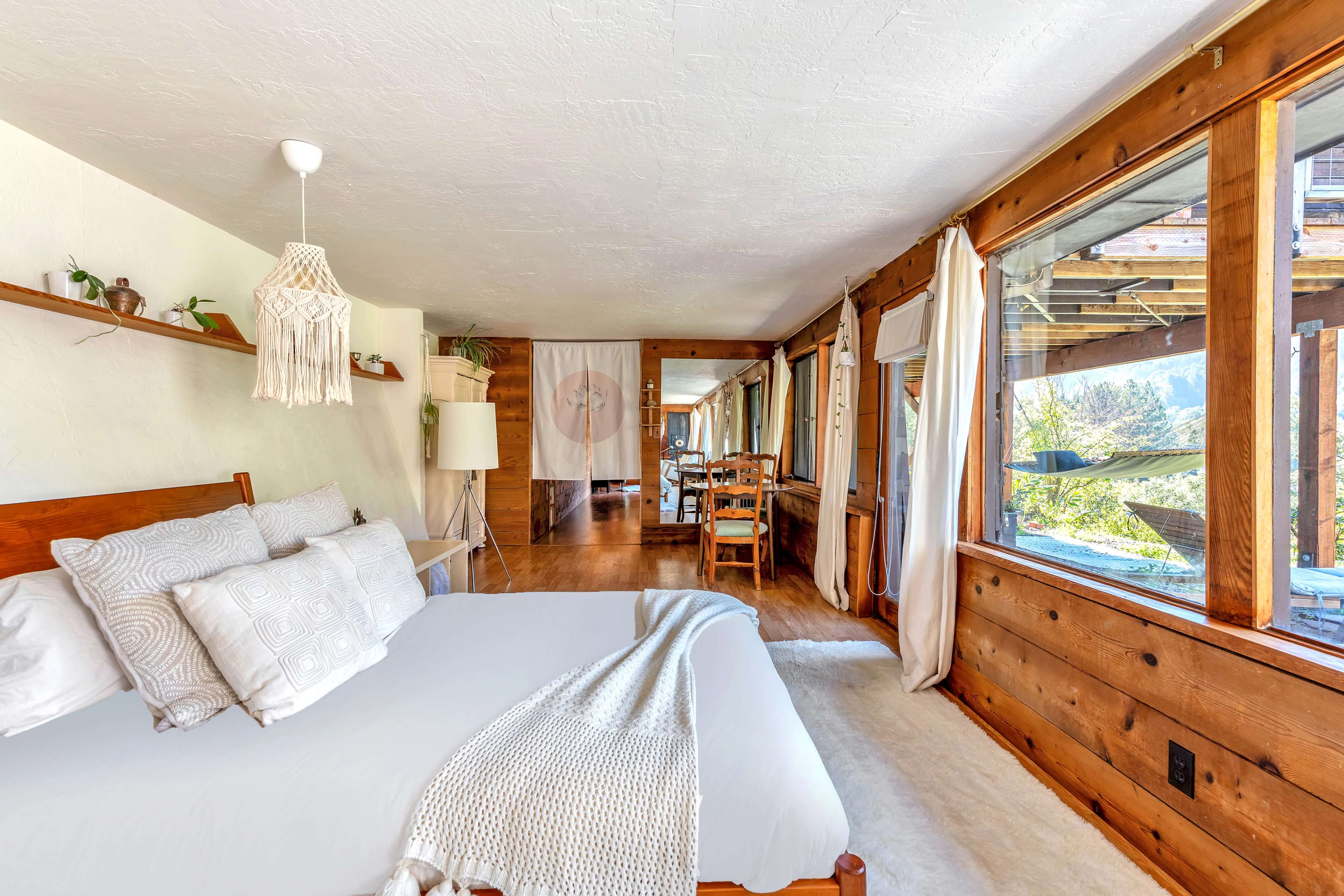 The image shows a bright and airy bedroom with a wooden bed, large windows, and a cozy seating area that leads into a dining space.