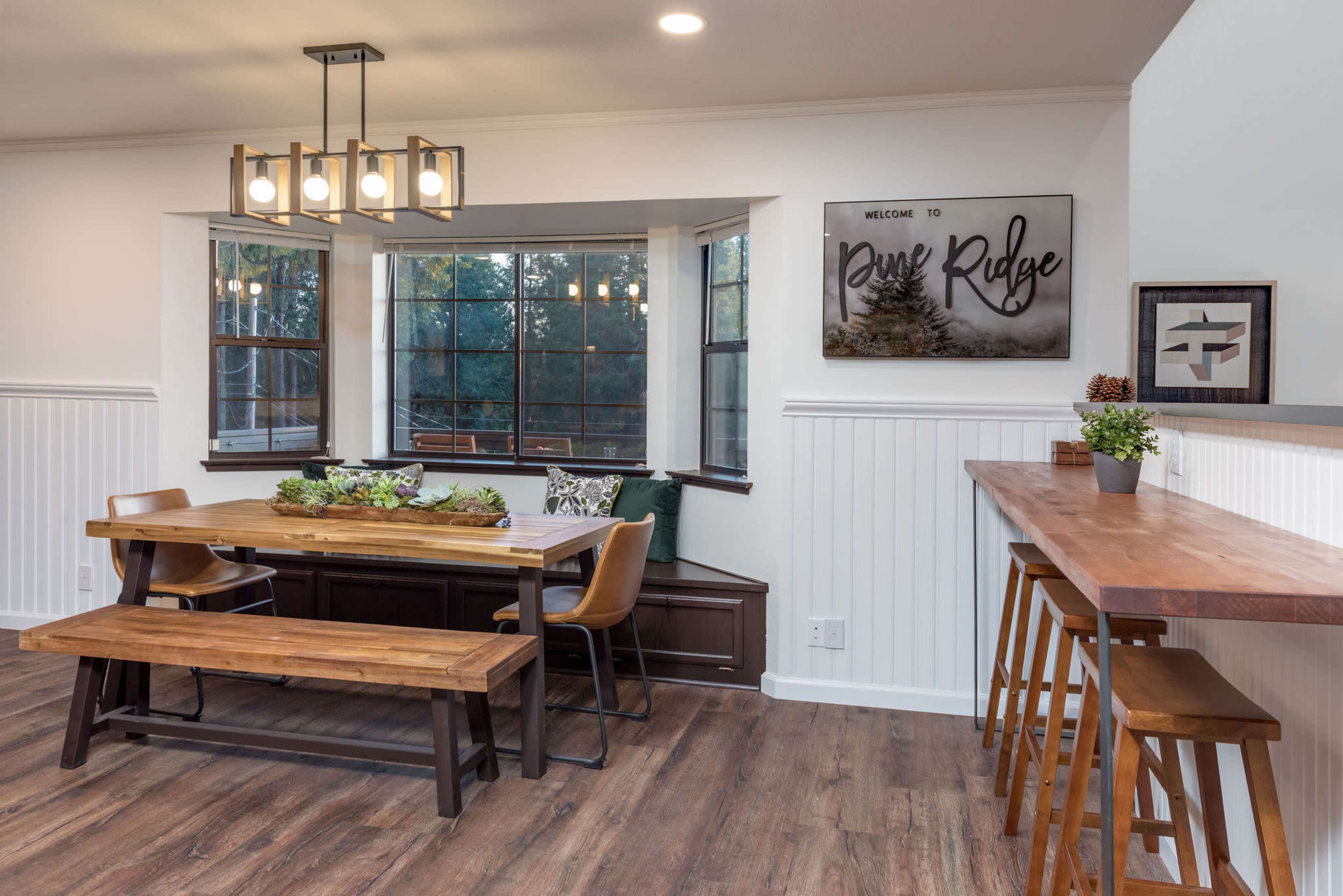 A cozy dining area featuring a wooden table with benches, a wall-mounted artwork, and large windows that allow natural light to fill the space.
