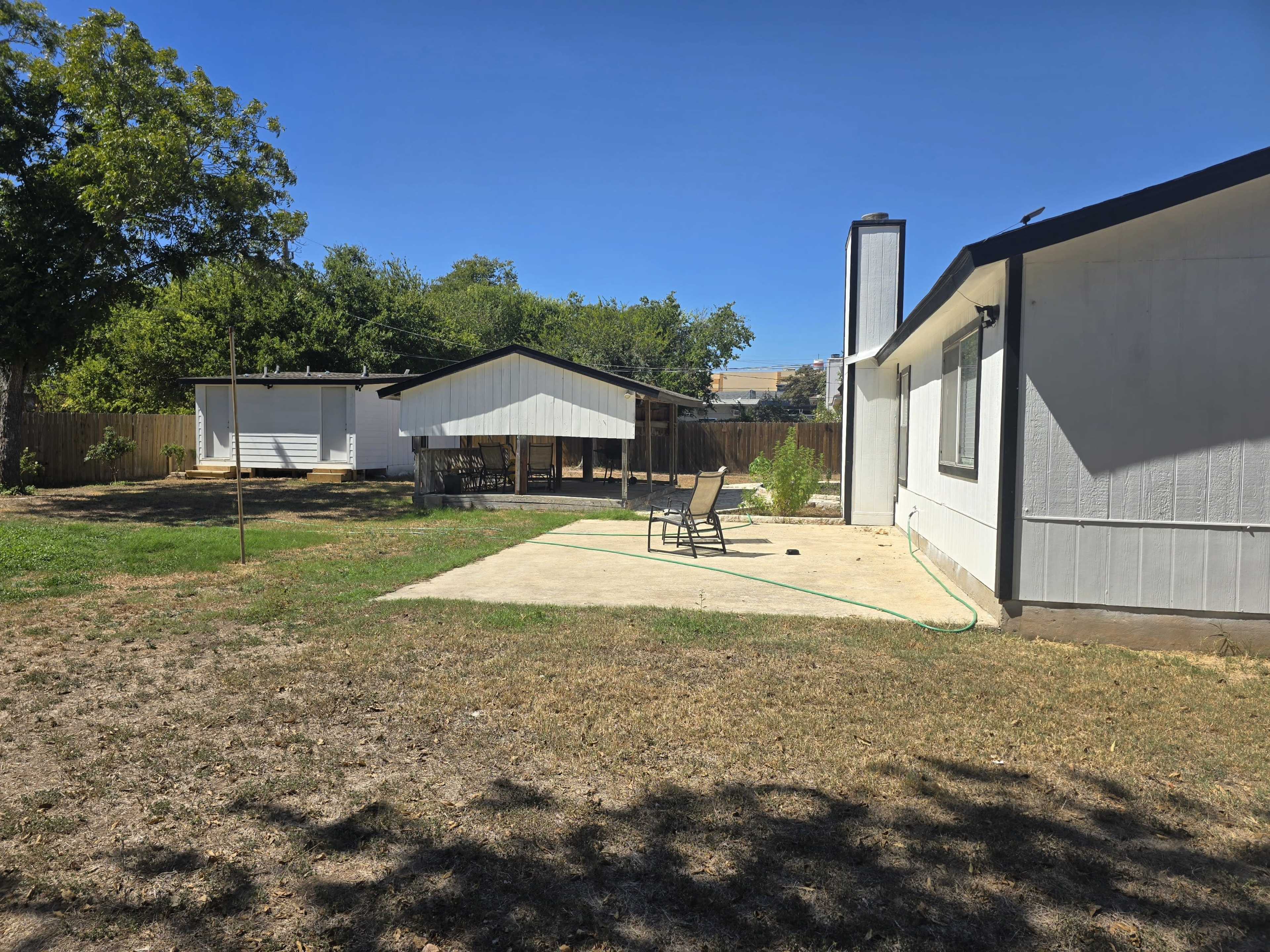 The image shows a backyard featuring a concrete patio with a chair, a white shed, and a single-story house under a clear blue sky.