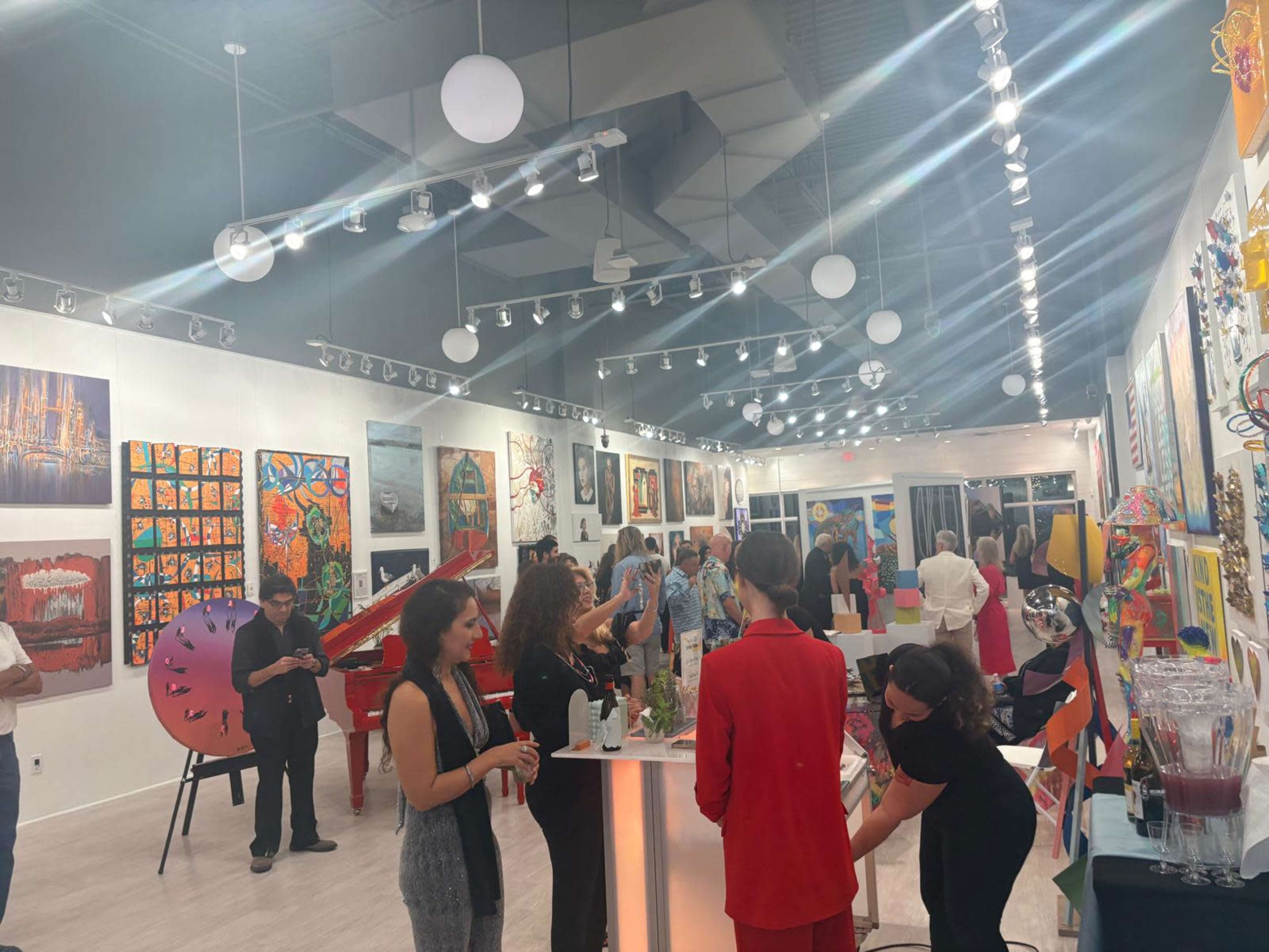 A group of people is gathered in an art gallery filled with various colorful artworks and a red grand piano.