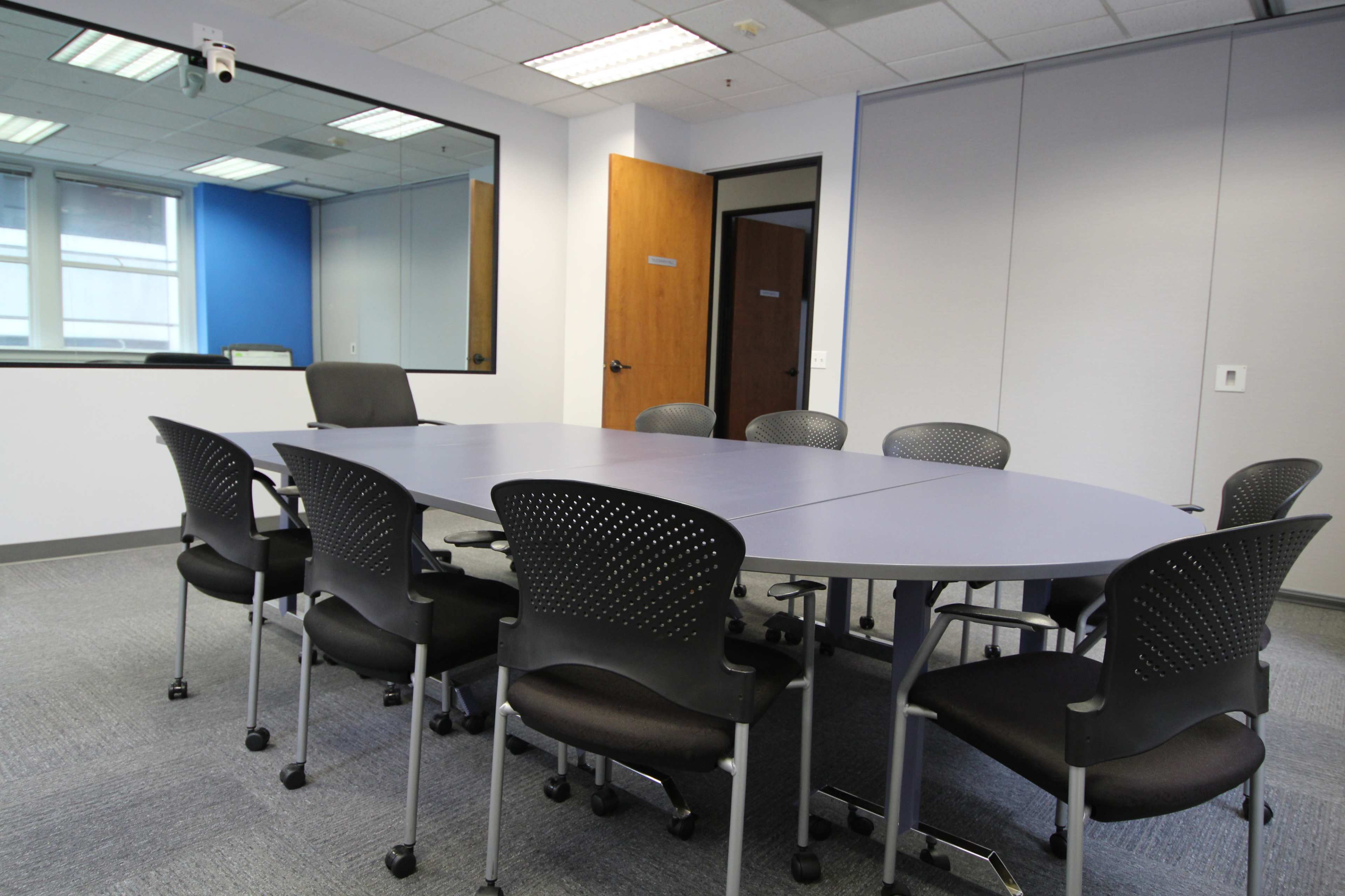A conference room features an oval table surrounded by eight black rolling chairs.