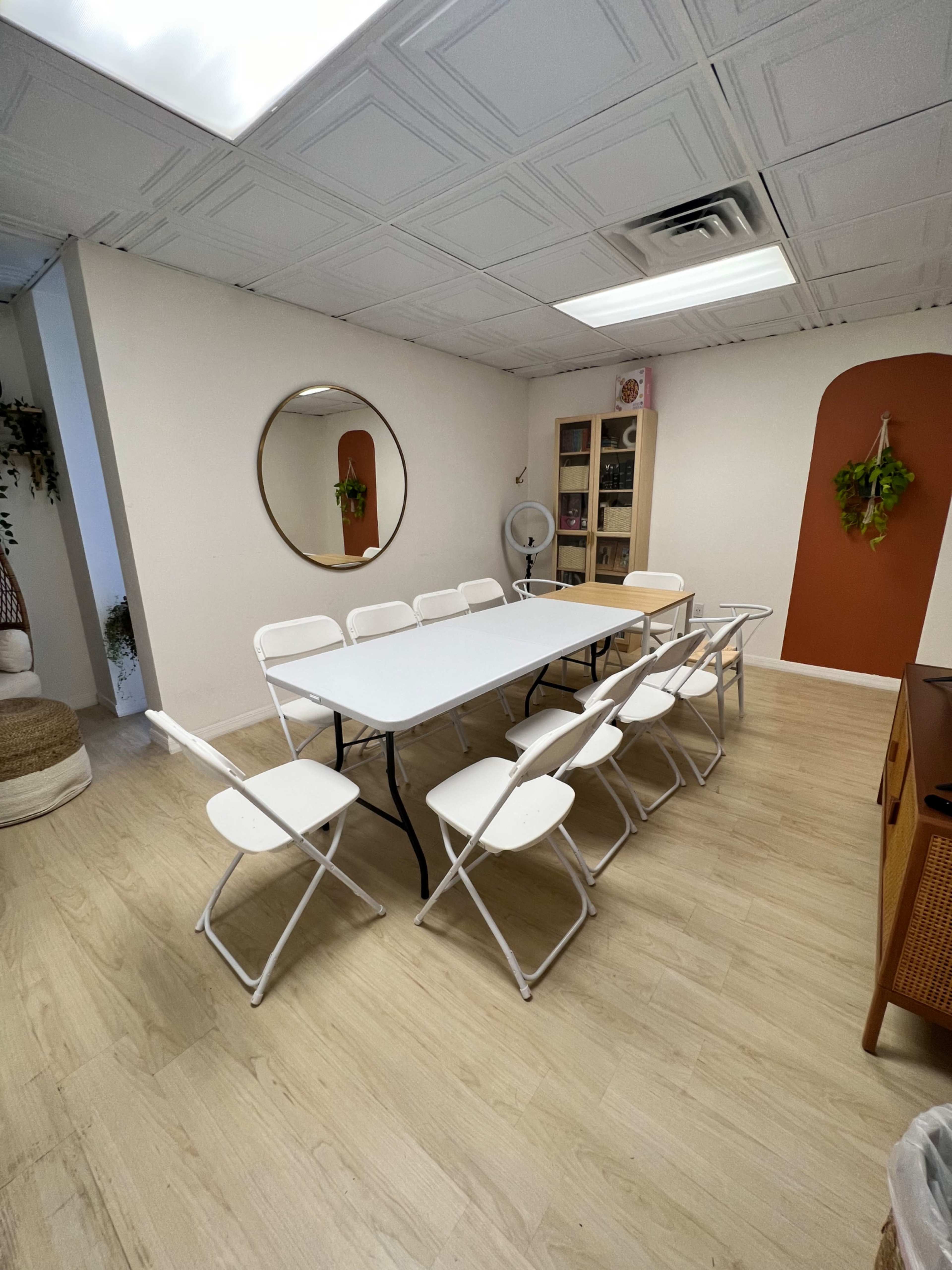A meeting room with a long white table surrounded by eight folding chairs, a large round mirror on the wall, and shelving containing various items.