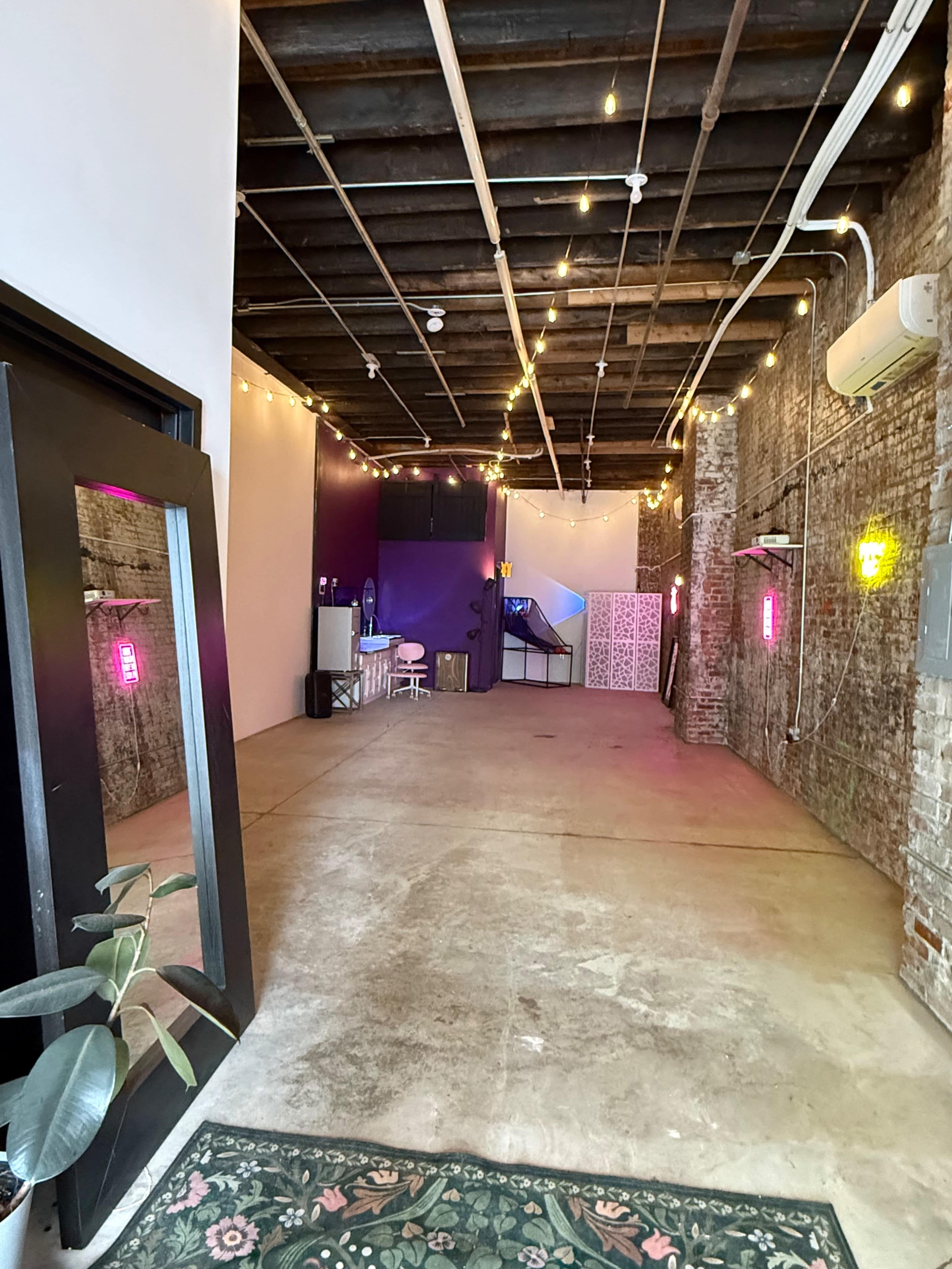 The image shows an interior space with exposed brick walls, a concrete floor, and string lights overhead, featuring a purple accent wall and a mirror at the entrance.