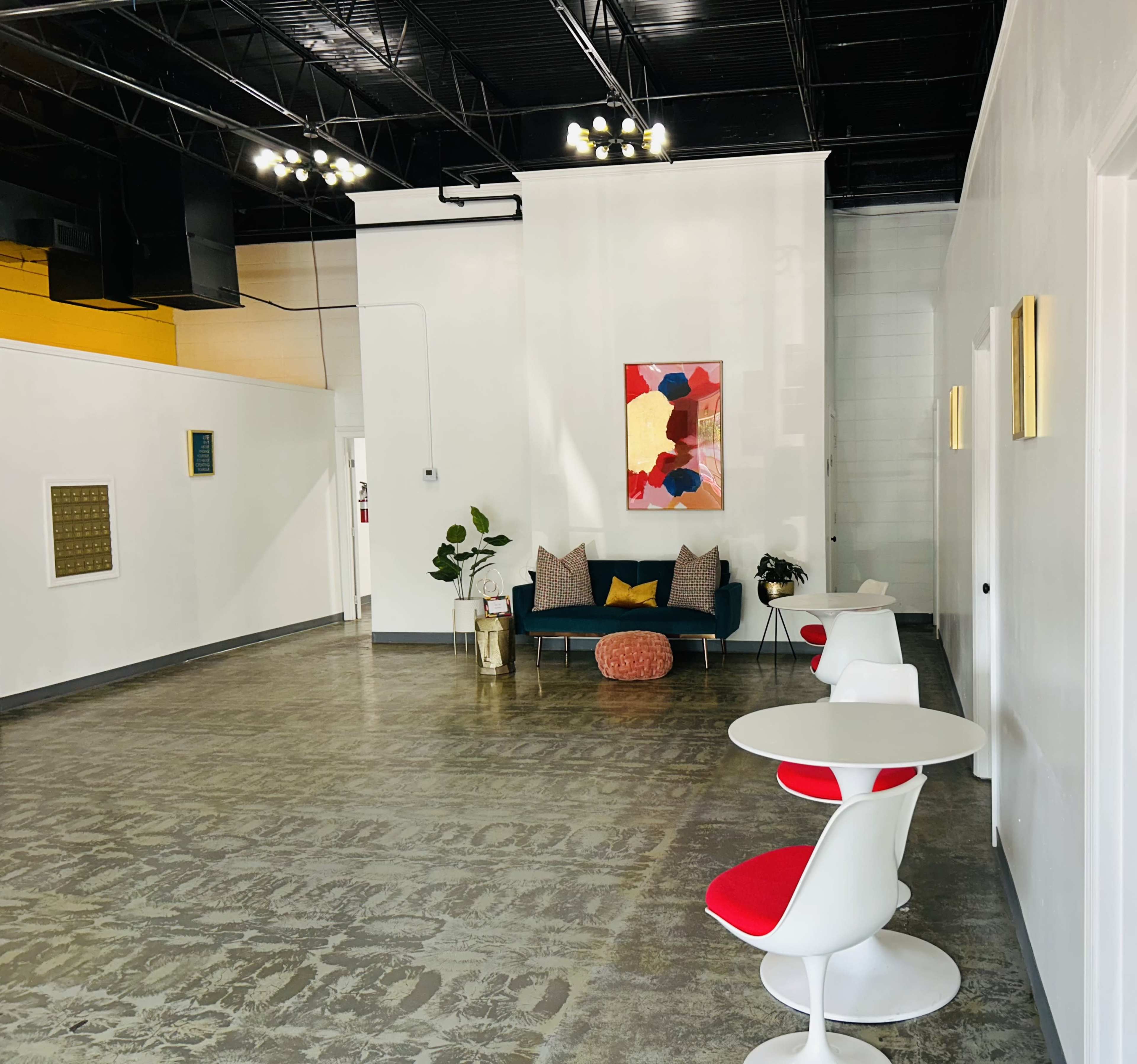 The image shows a spacious, modern interior with white walls, a colorful abstract painting, a blue couch, and minimalist round tables with red accents.