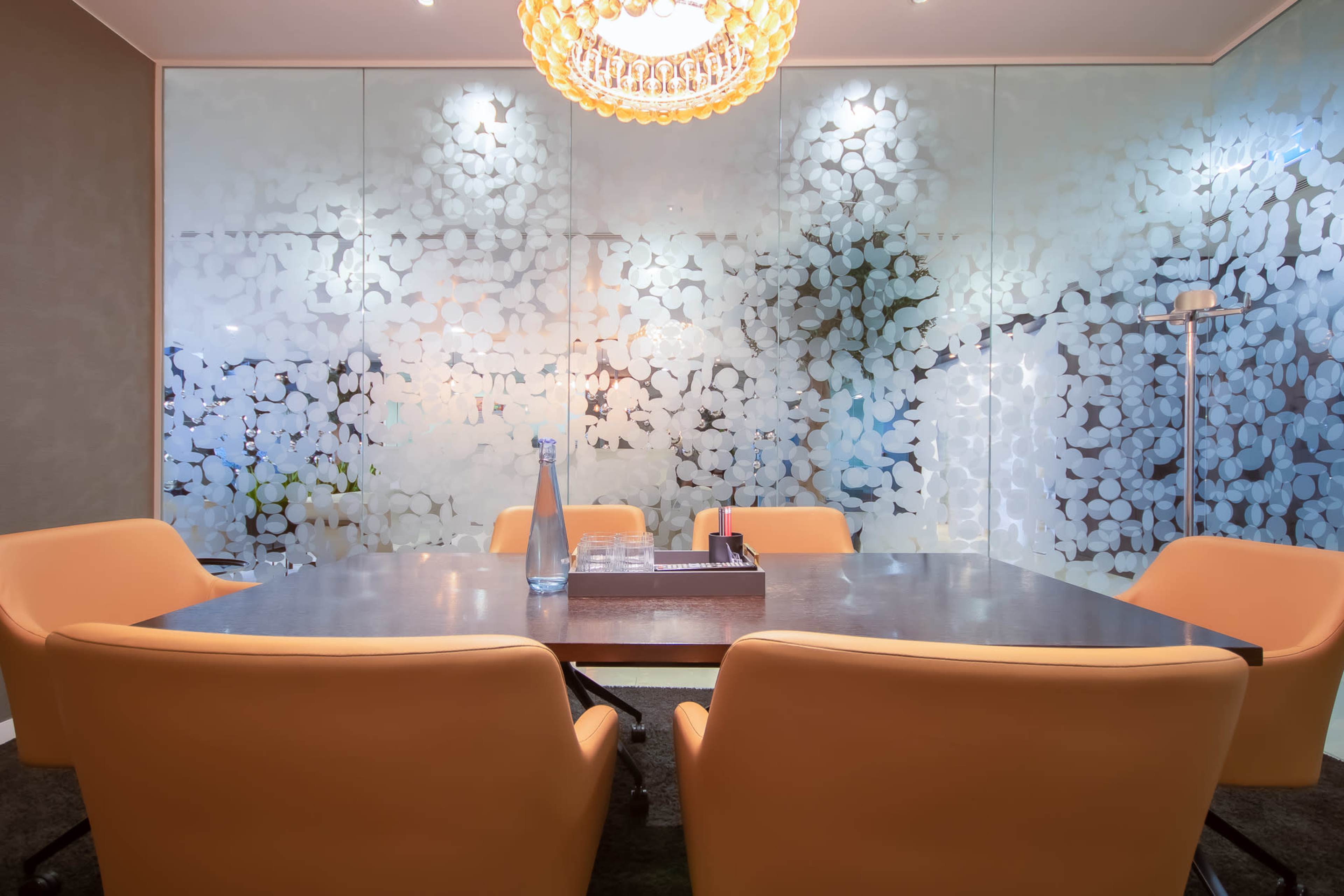 A modern conference room features a large table surrounded by four orange chairs and a decorative glass wall.