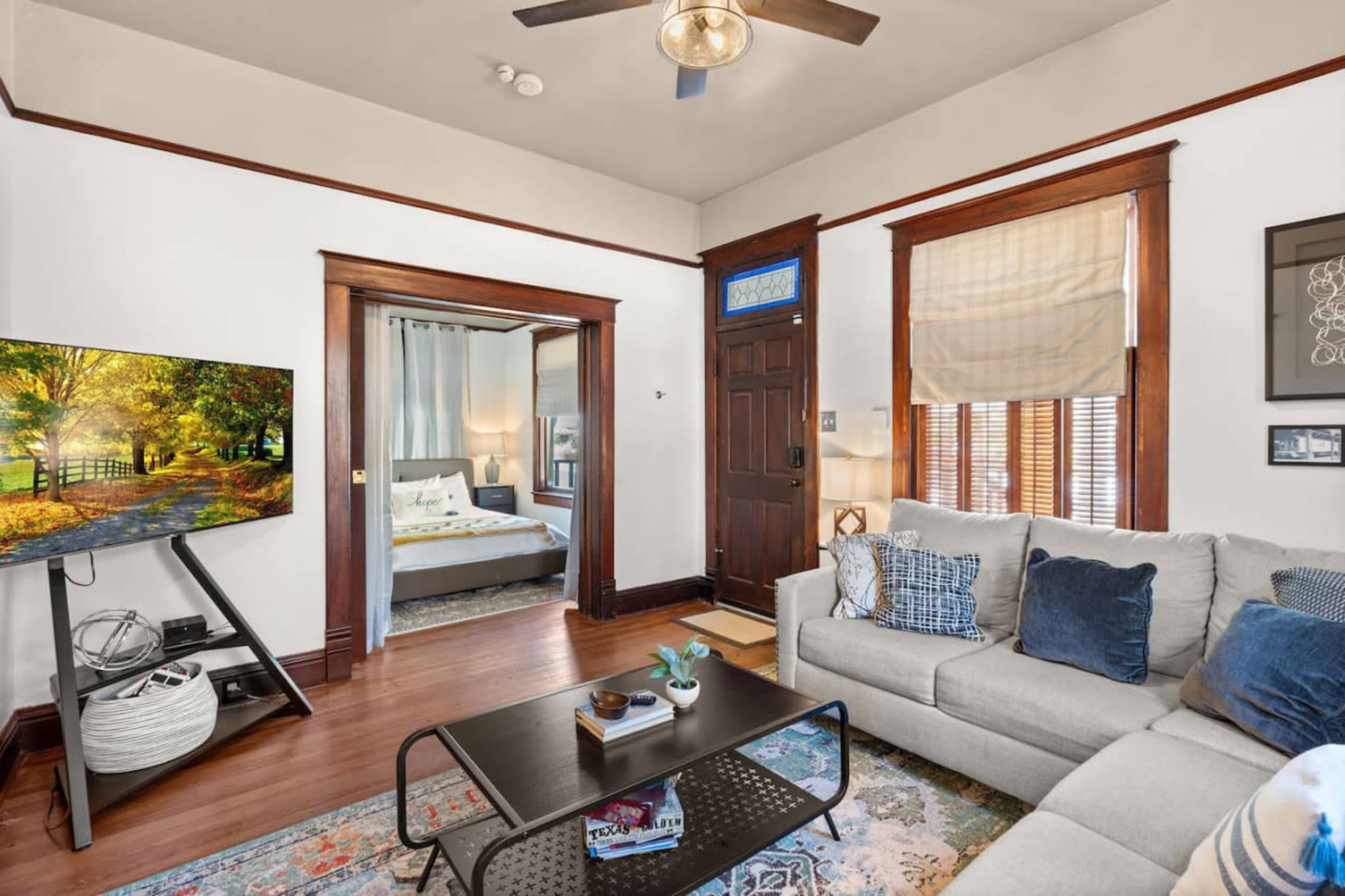 The living room features a light gray couch, a coffee table on a patterned rug, a television on a stand, and a doorway leading to a bedroom with a large window.
