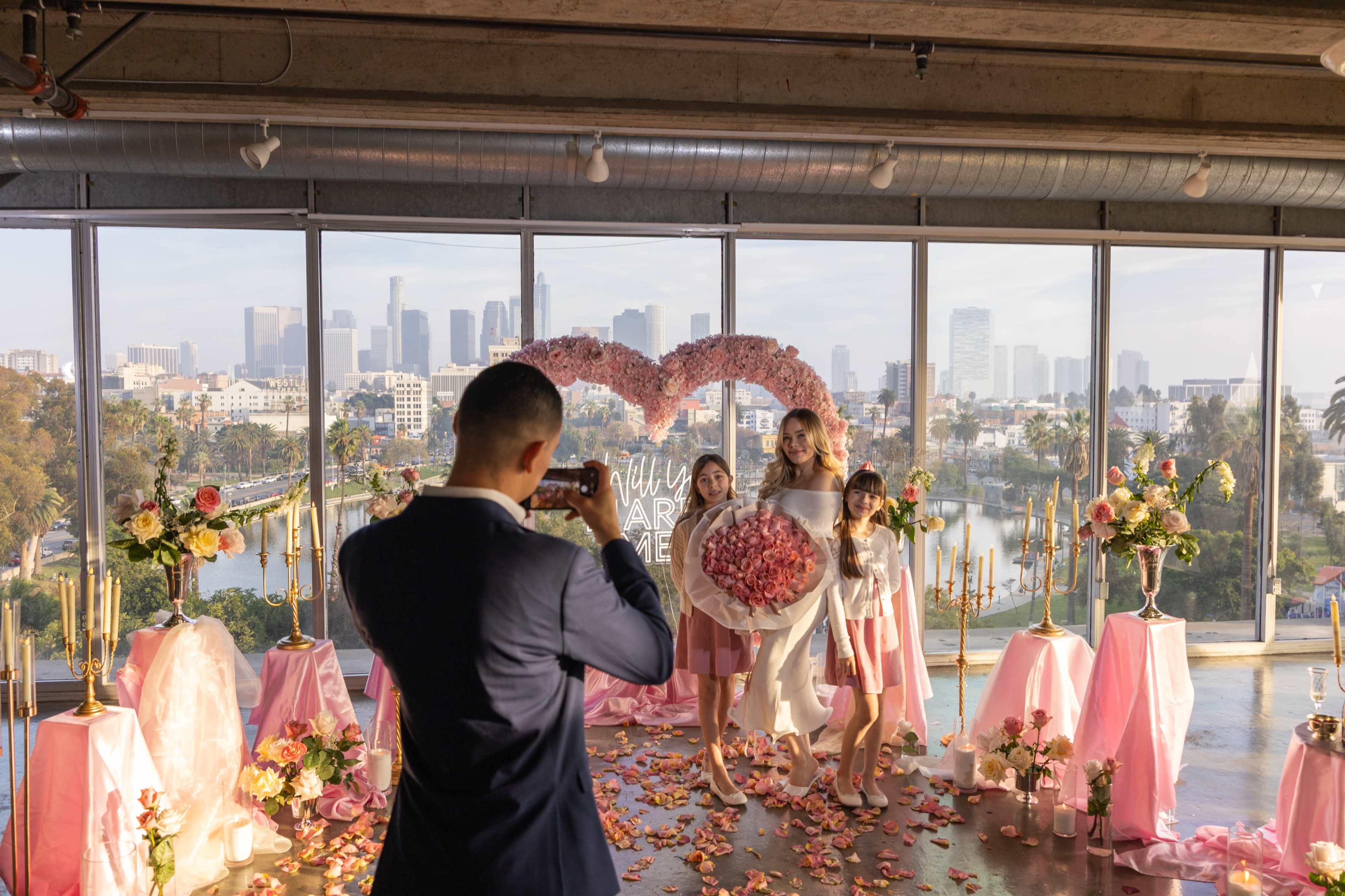 A man takes a photograph of a woman and two girls standing in front of a floral heart backdrop during a celebration in a city view venue.