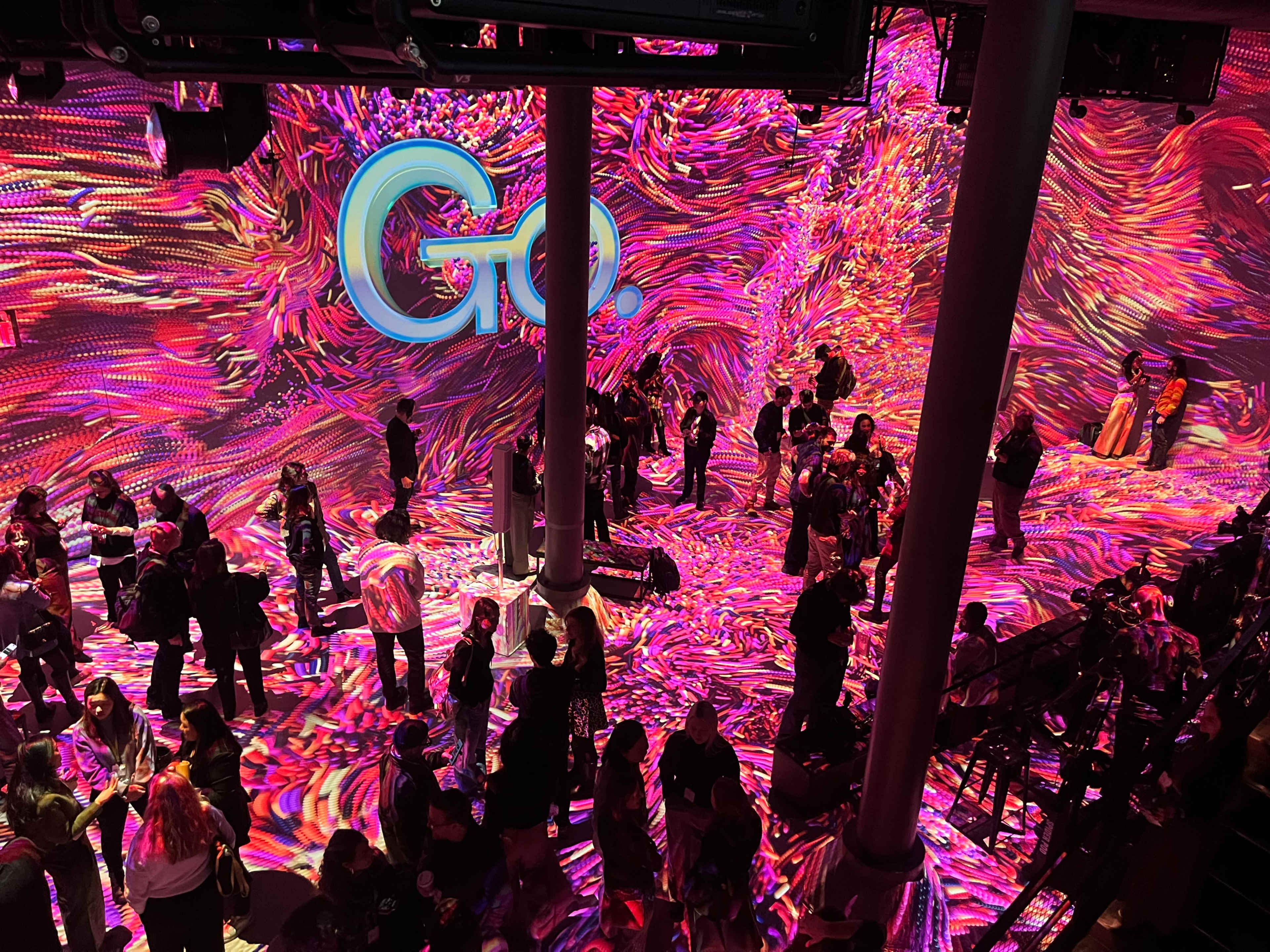 A vibrant, colorful digital installation fills the space with swirling patterns, while people gather and interact throughout the venue.