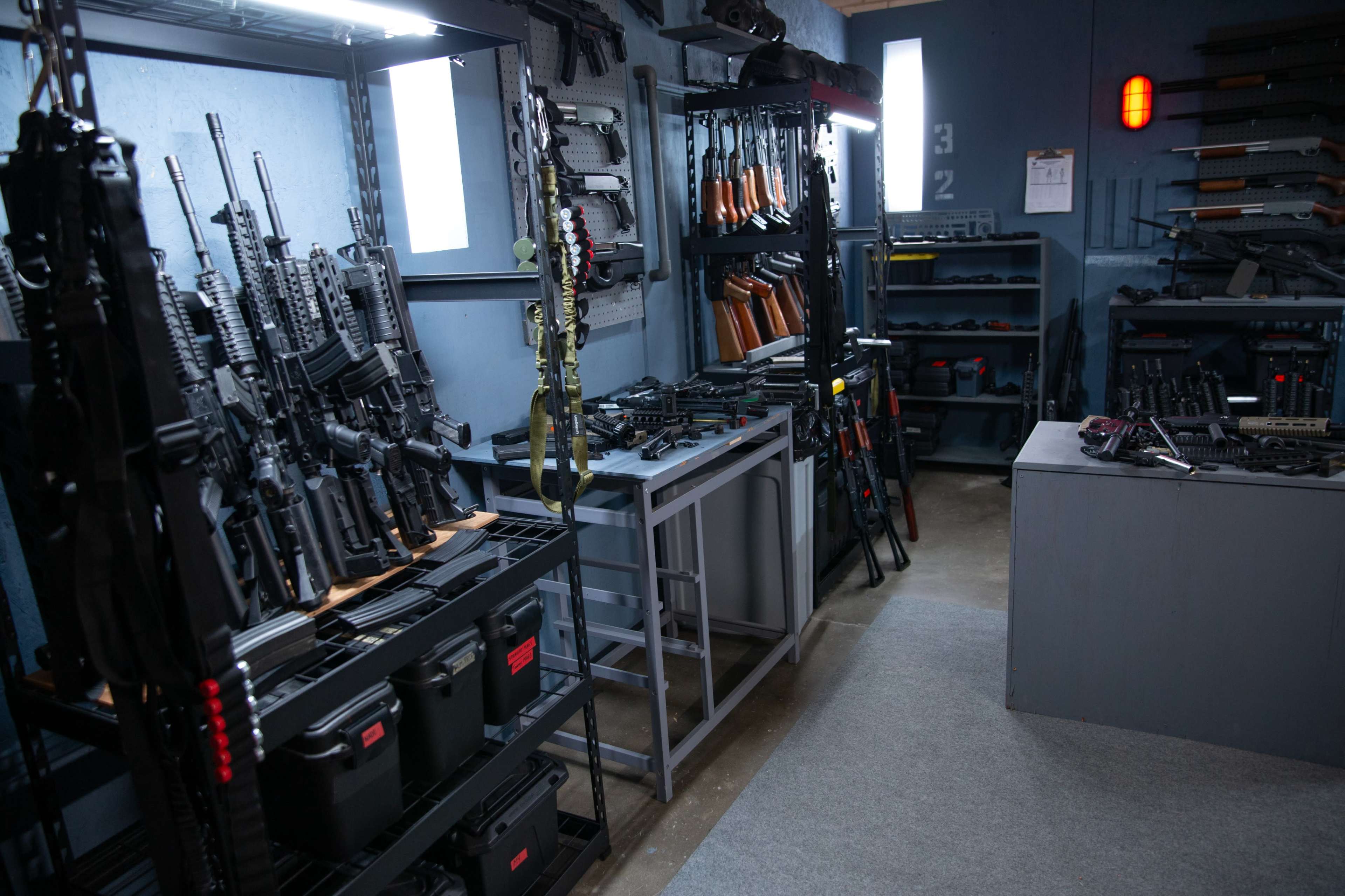 The image shows a room filled with various firearms displayed on racks and shelves, with a focus on organized weaponry and storage boxes.