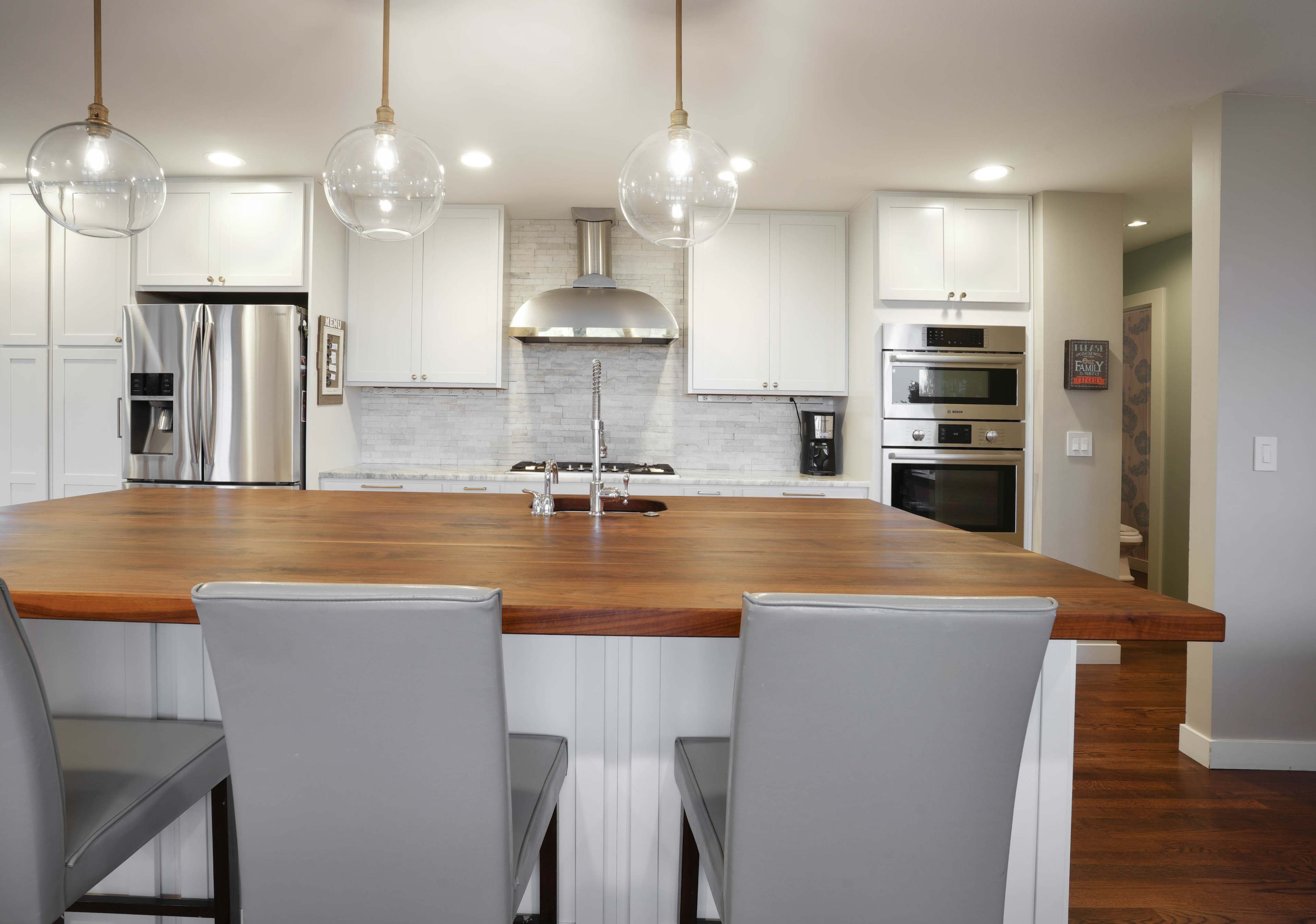 A modern kitchen features a large wooden island with gray bar stools, stainless steel appliances, and white cabinetry.