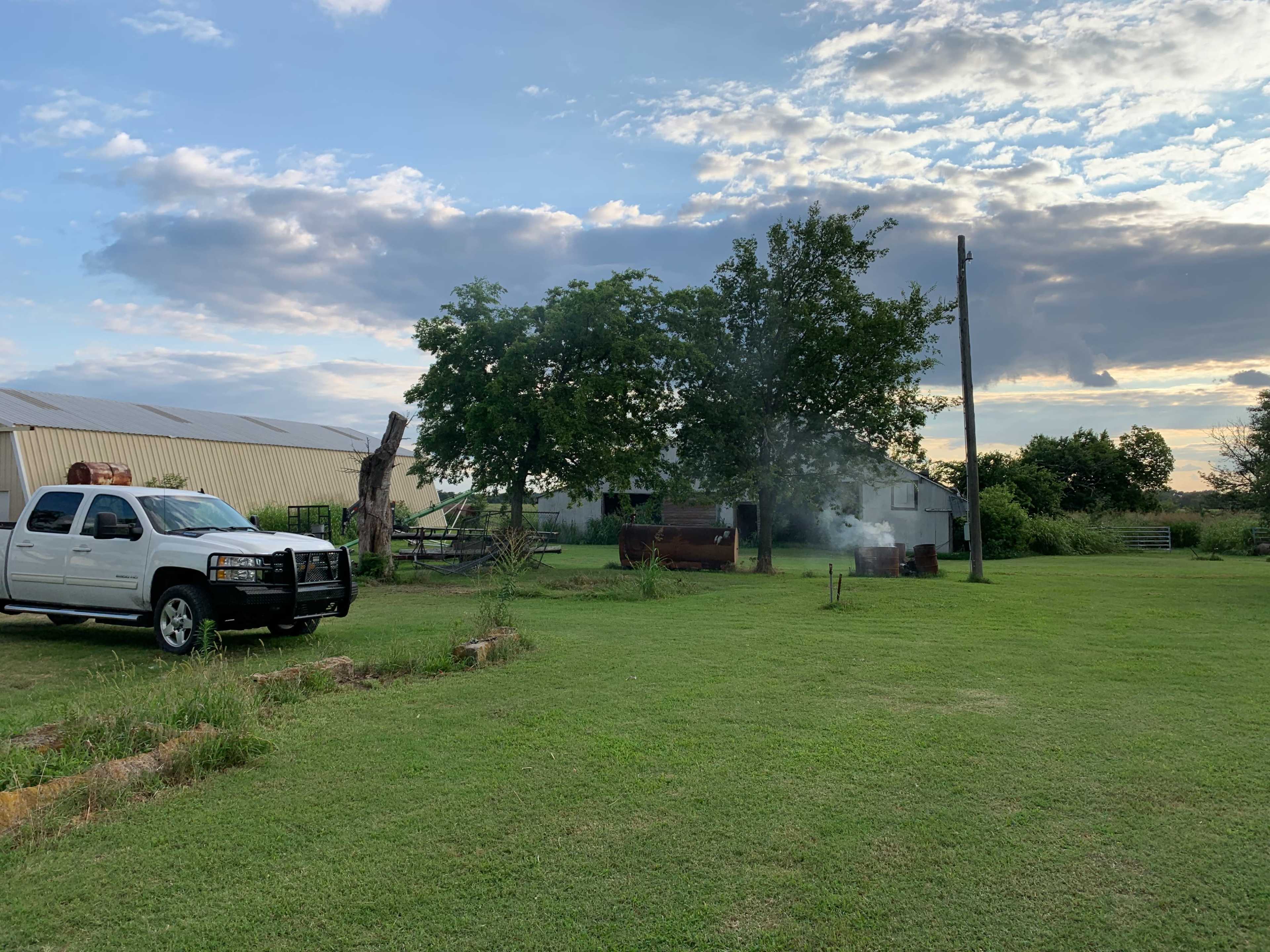 A white pickup truck is parked on a grassy area near a barn, with smoke rising from a nearby fire pit and a large tree in the background.