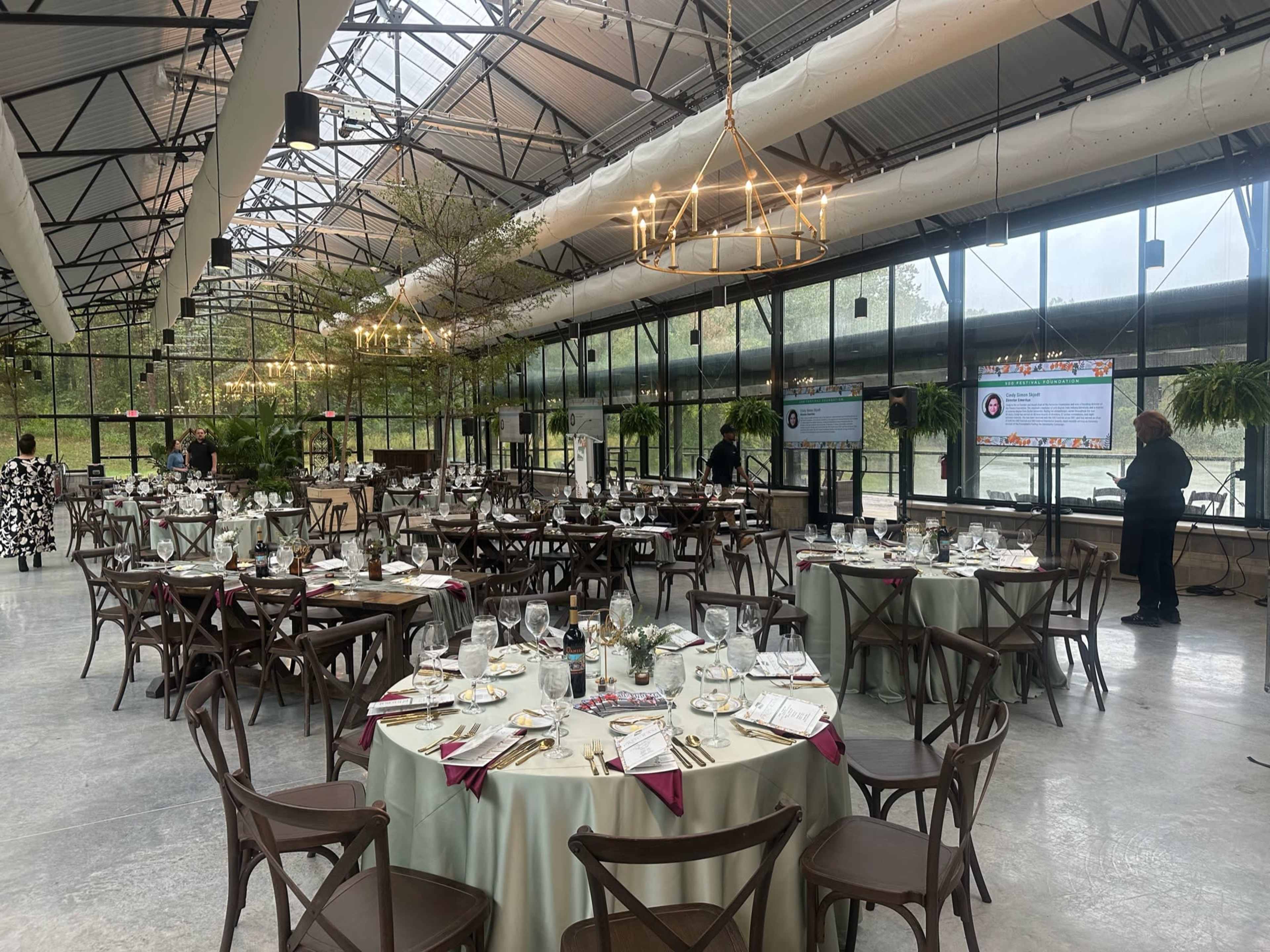 The image shows a spacious event venue with round tables set for dining, surrounded by large windows and hanging plants.