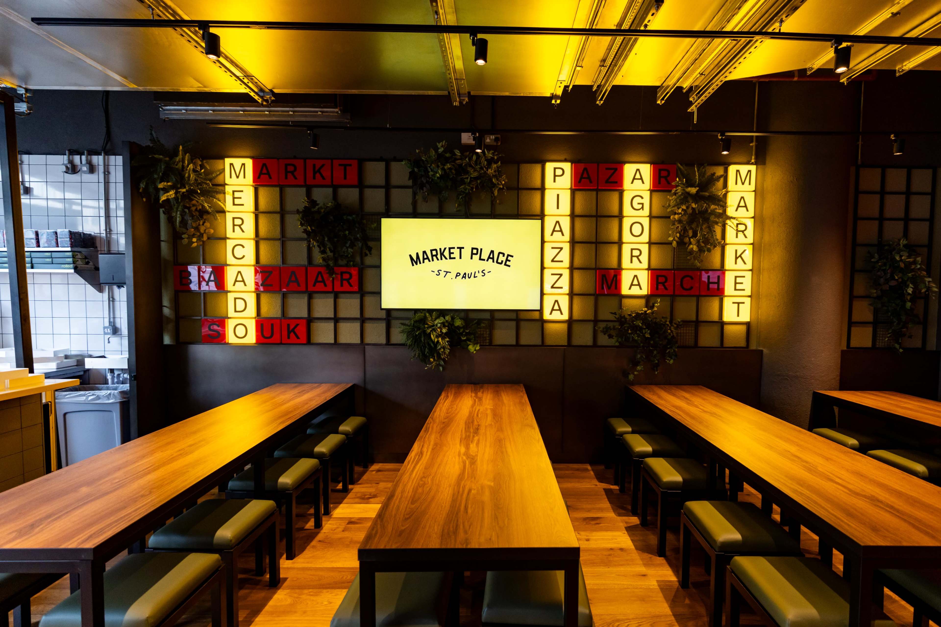 The image shows a modern dining space with long wooden tables, green benches, and a large screen displaying the words "MARKET PLACE ST. PAUL'S" surrounded by colorful letter lights.