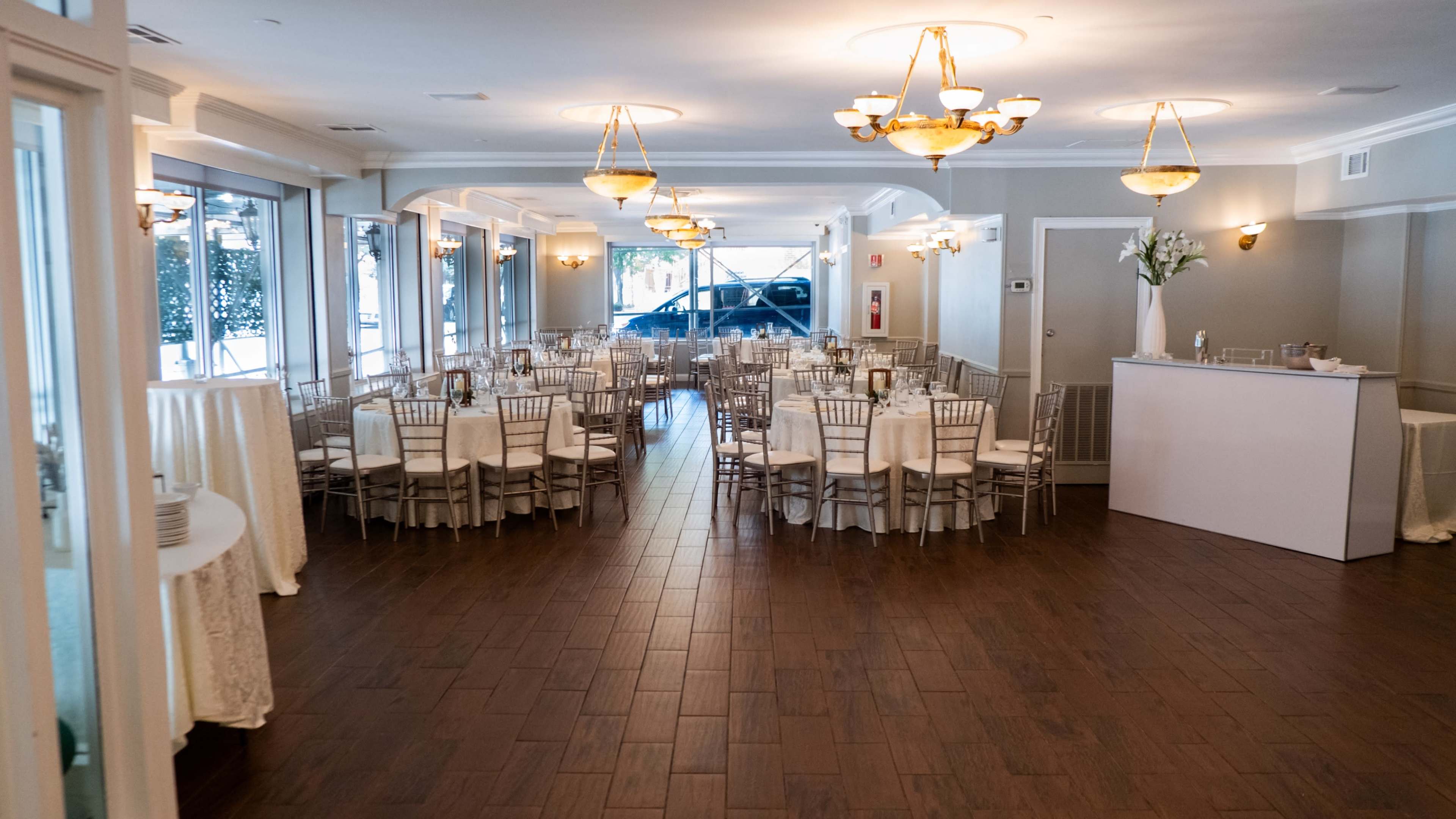 The image shows a banquet hall set up with rows of tables and chairs, a bar area, and large windows allowing natural light to enter.