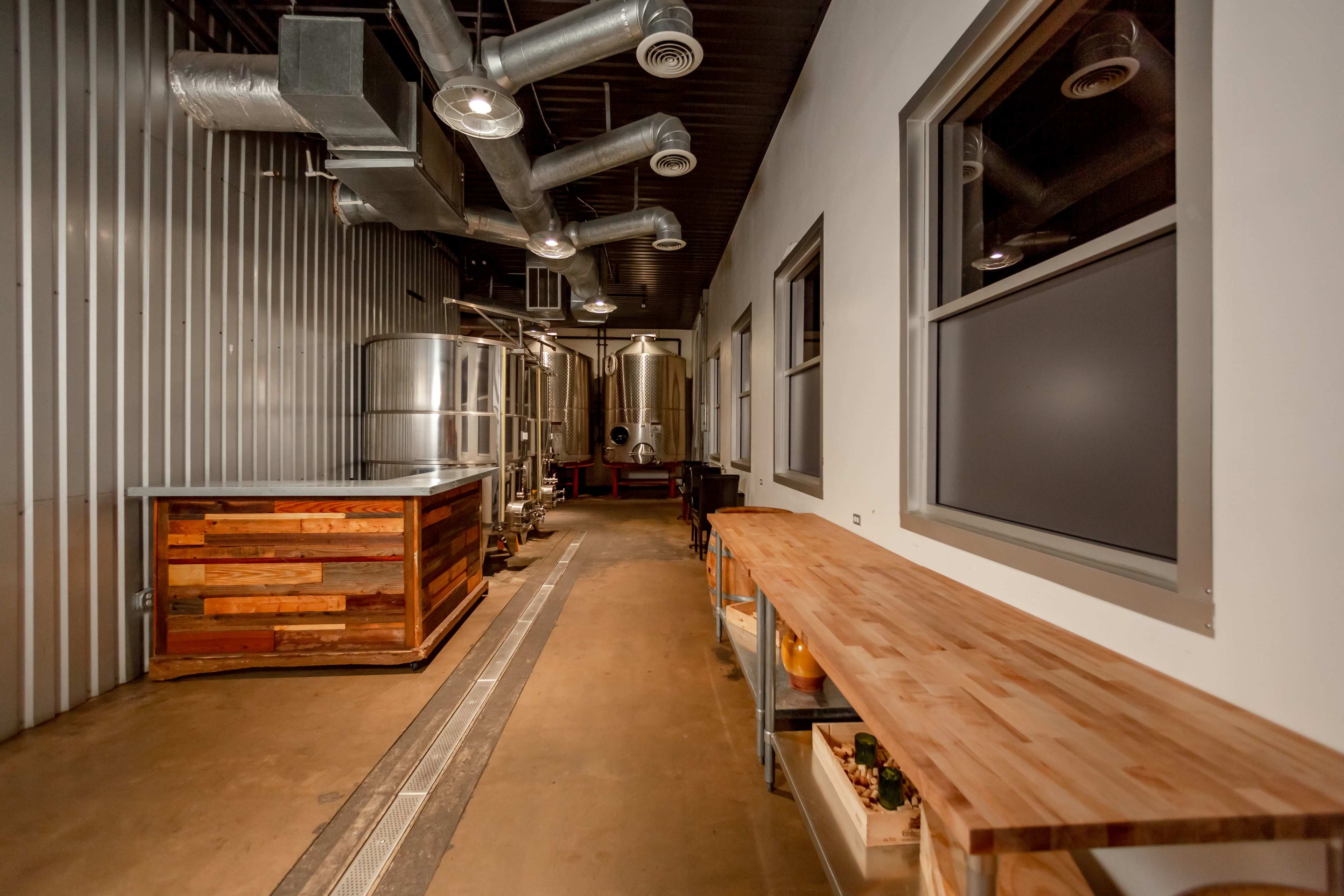 The image shows a narrow industrial space with large metal brewing tanks, a wooden countertop, and exposed ductwork along the ceiling.