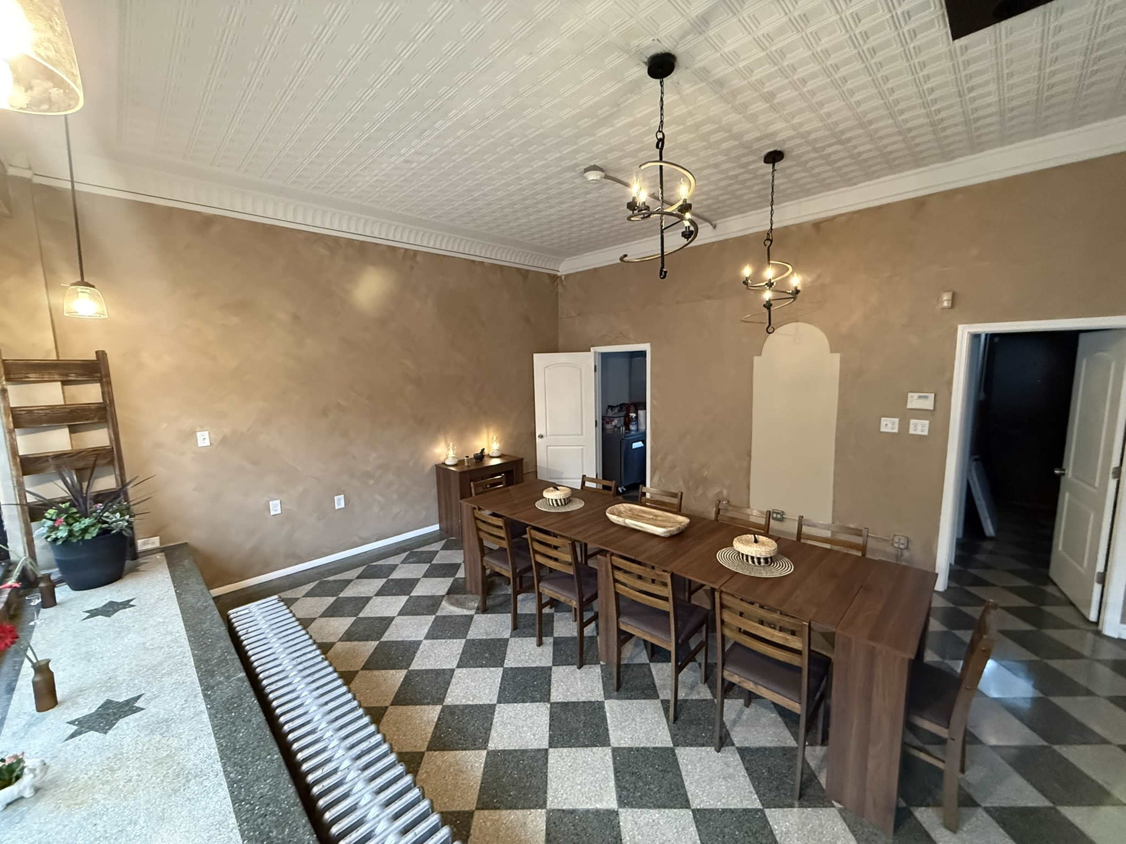 The image shows a spacious dining area with a large wooden table surrounded by eight chairs, set against a backdrop of textured beige walls and a checkered floor.