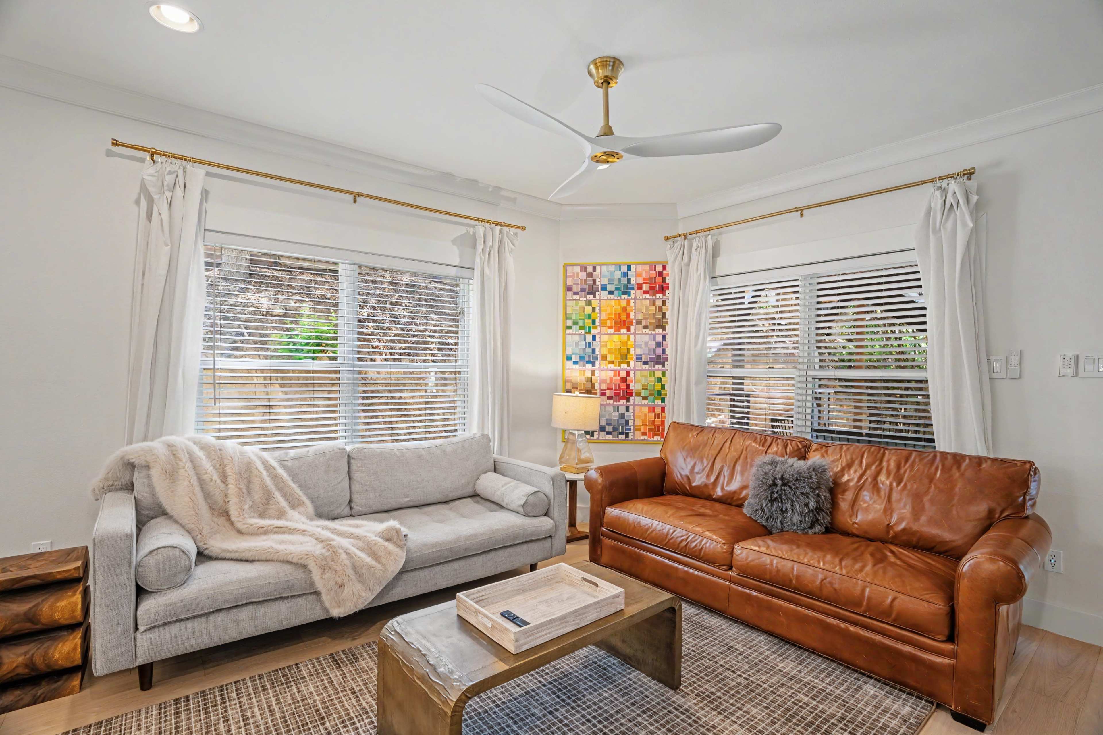The image shows a living room with two sofas, a coffee table, a lamp, and colorful wall art, all illuminated by natural light from large windows.