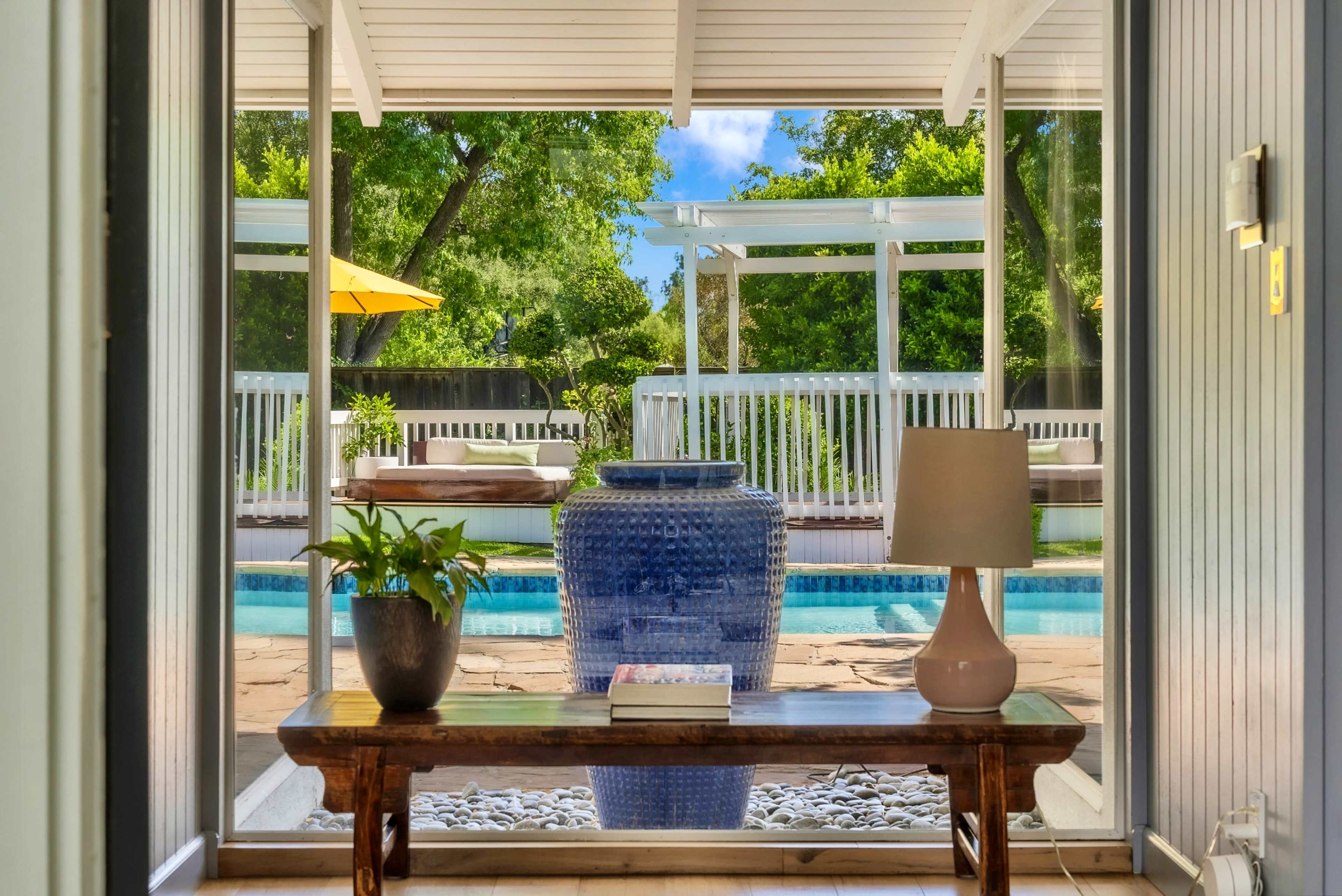 The image shows a view from an indoor space featuring a table with a lamp and a potted plant, overlooking a swimming pool and patio area through large glass doors.