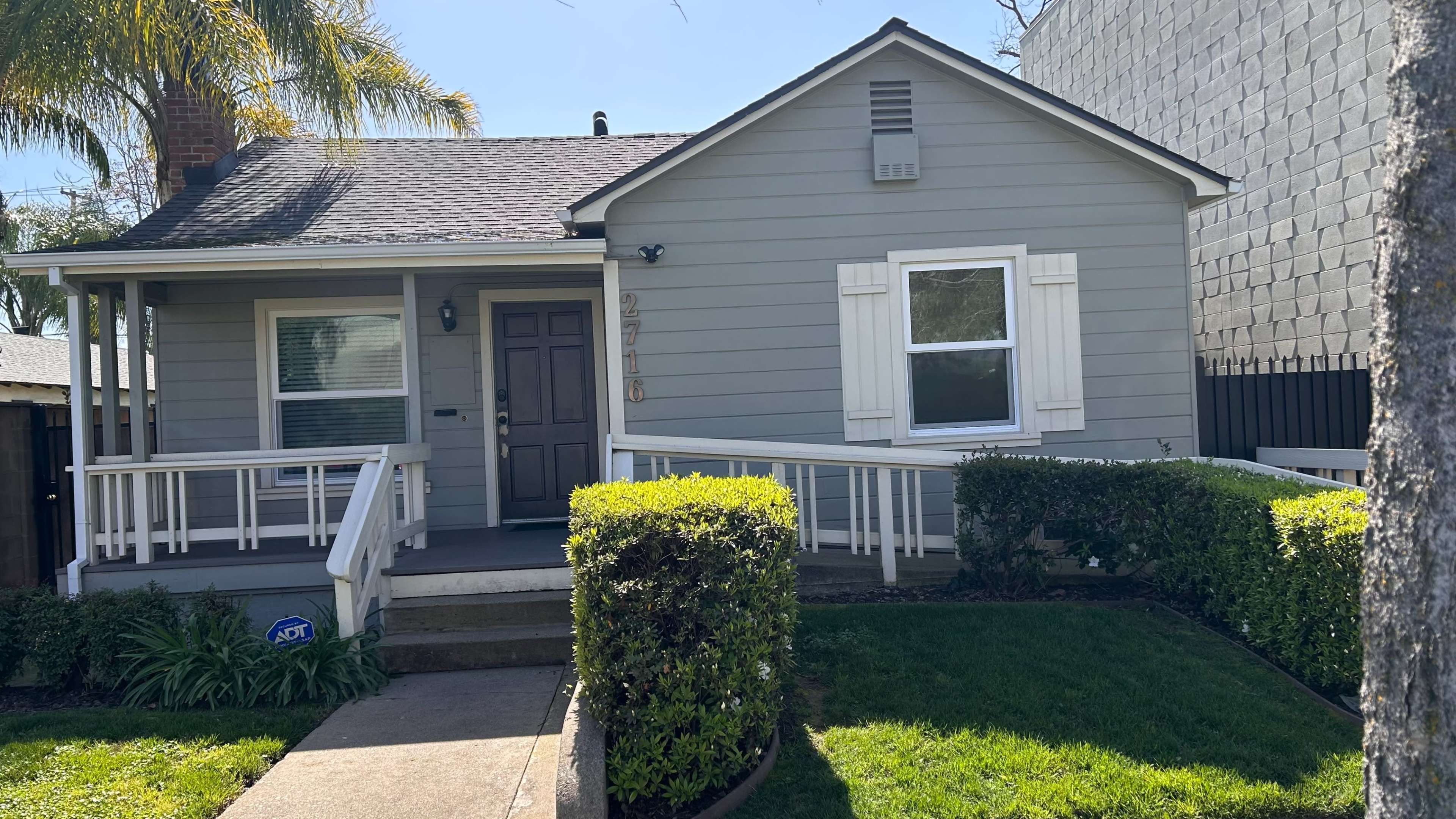A small gray house with white shutters and a front porch is surrounded by neatly trimmed hedges and grass.