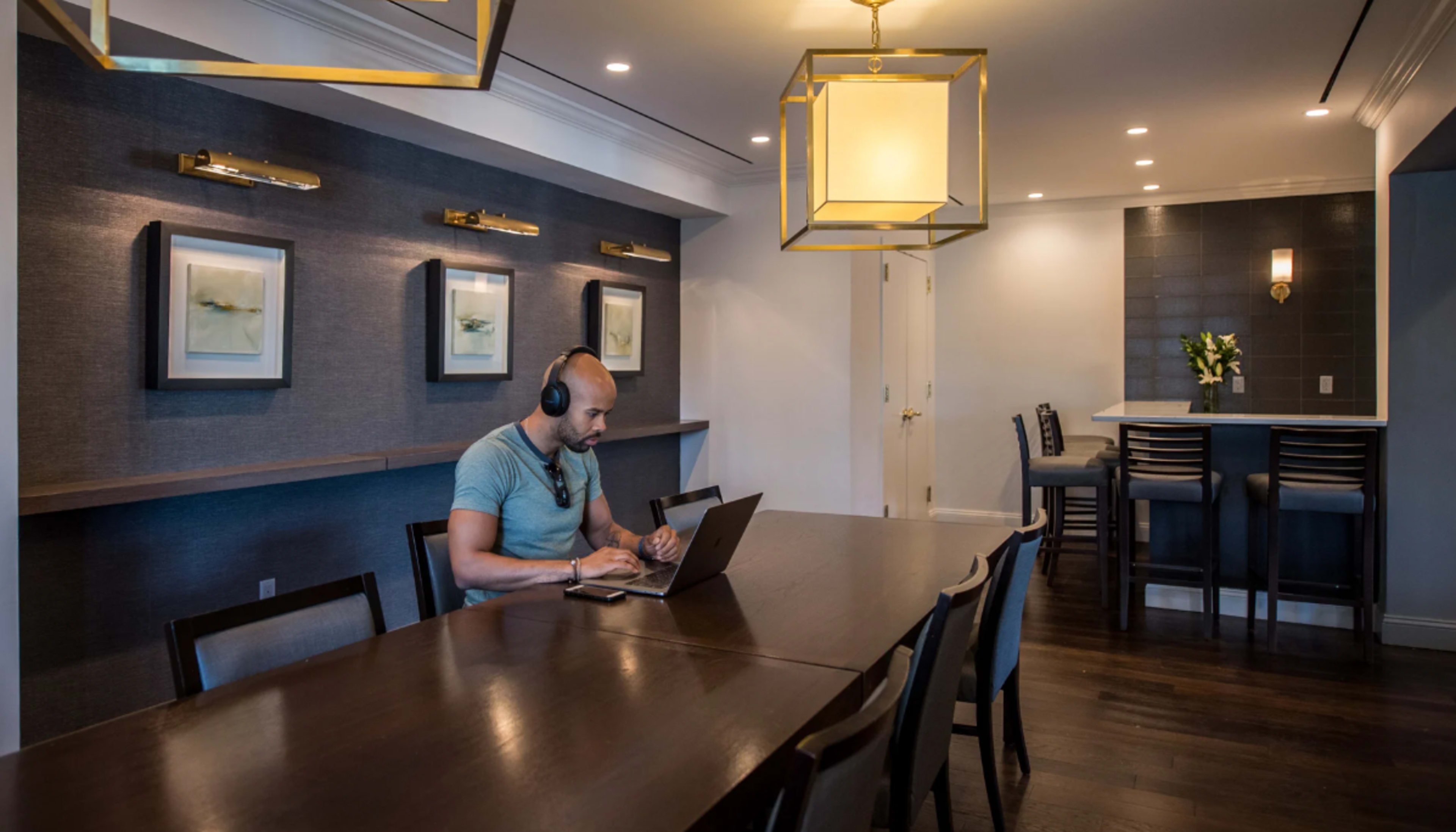 A man in headphones is sitting at a long dining table, working on a laptop in a well-lit room featuring framed artwork on the walls.