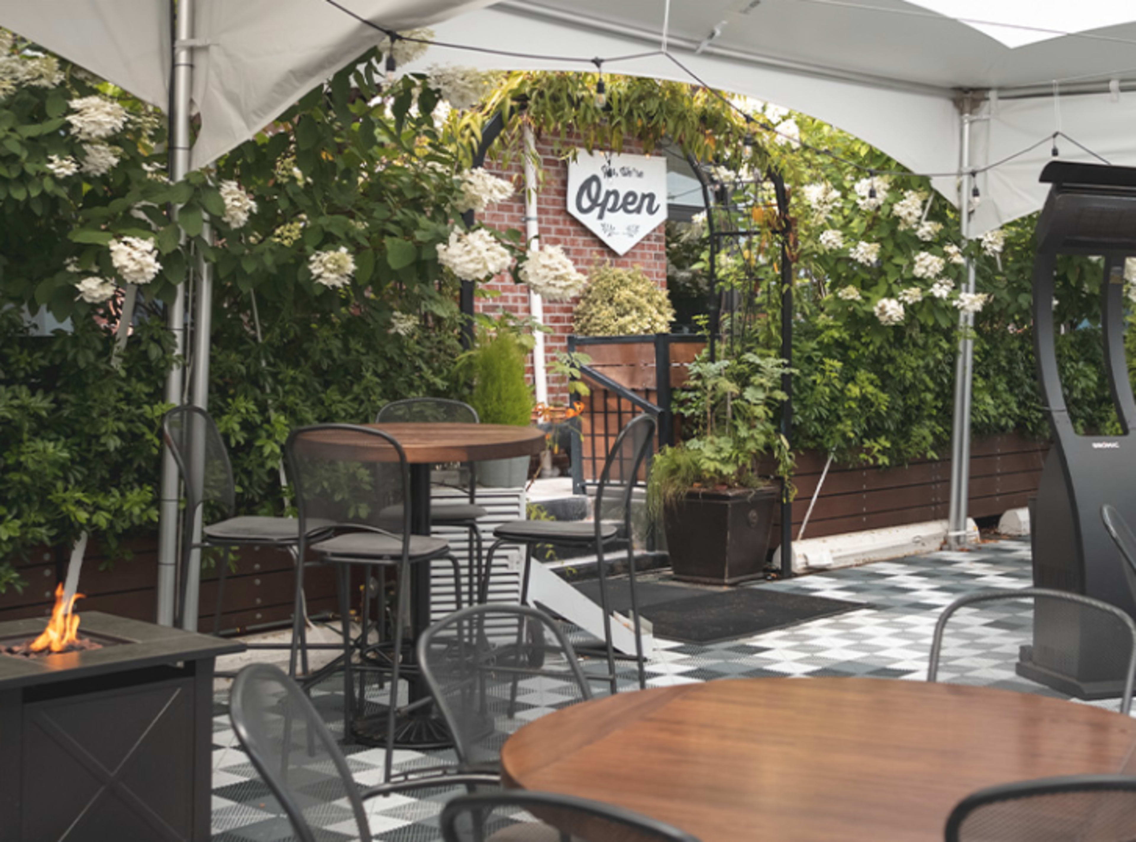 An outdoor seating area under a white tent, featuring several round tables and chairs, with lush greenery and a sign indicating that the space is open.